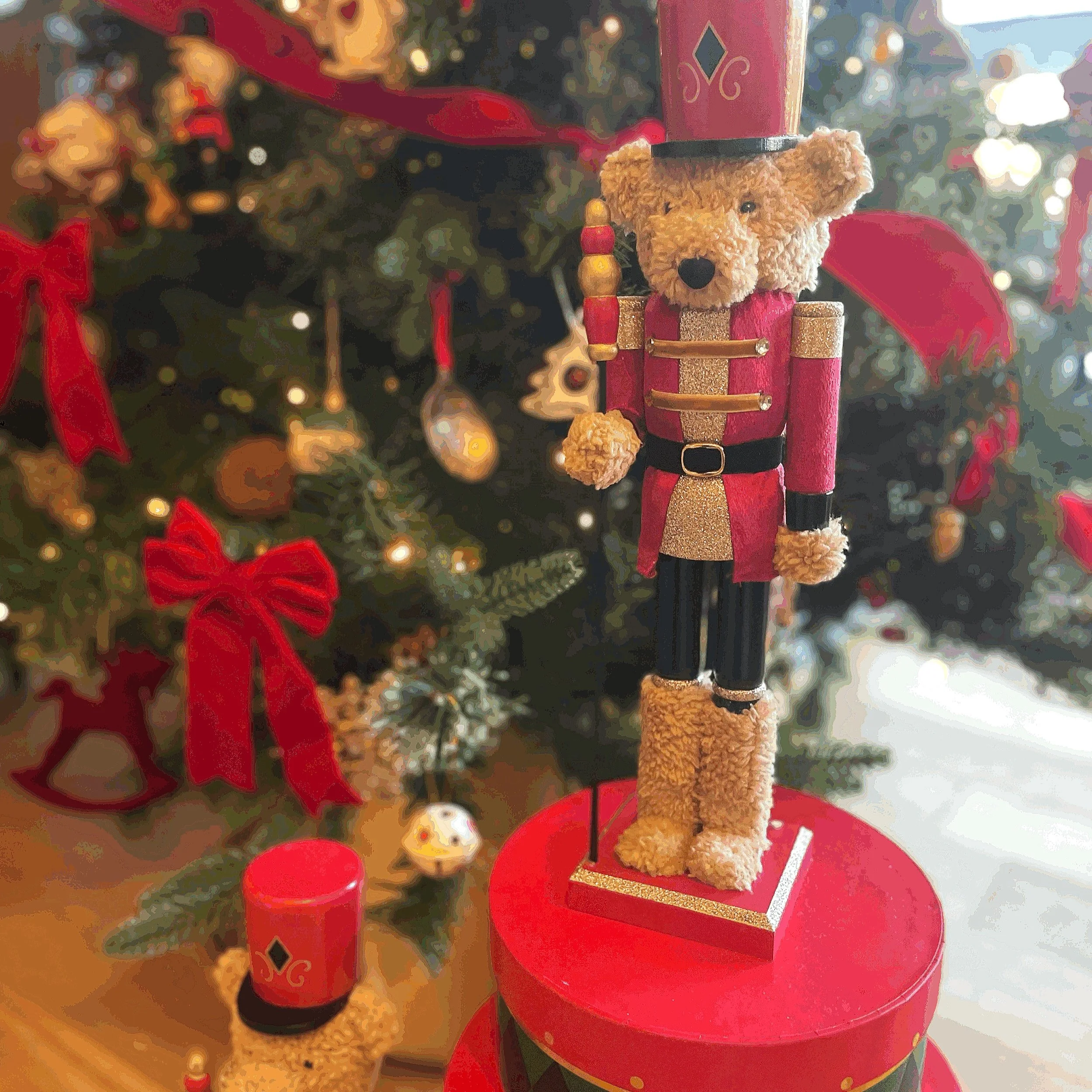 A decorative Christmas nutcracker with a teddy bear face, dressed as Santa Claus, standing next to a Christmas tree decorated with red ribbons and ornaments.