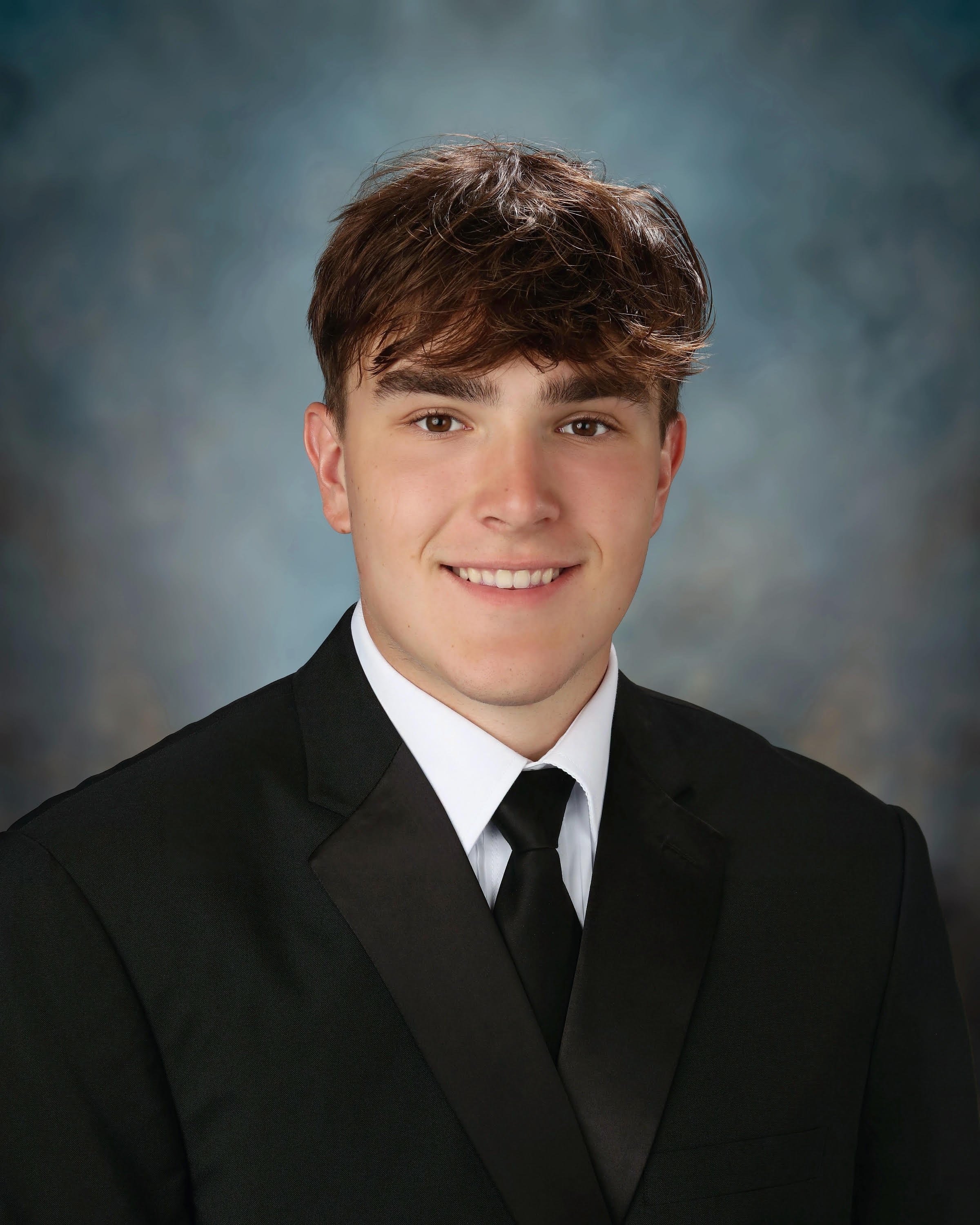 Close-up of a young man in a black tuxedo with a white shirt and black tie, smiling against a neutral background.