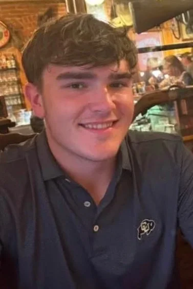 A young man with dark hair smiling, wearing a dark polo shirt, sitting in a restaurant or bar with a brick wall background and other patrons.