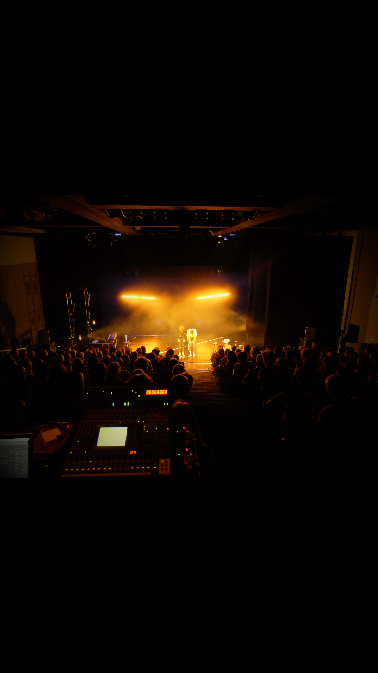 Une scène de concert avec des artistes sur scène, éclairage orange, et un public assis regardant la performance.