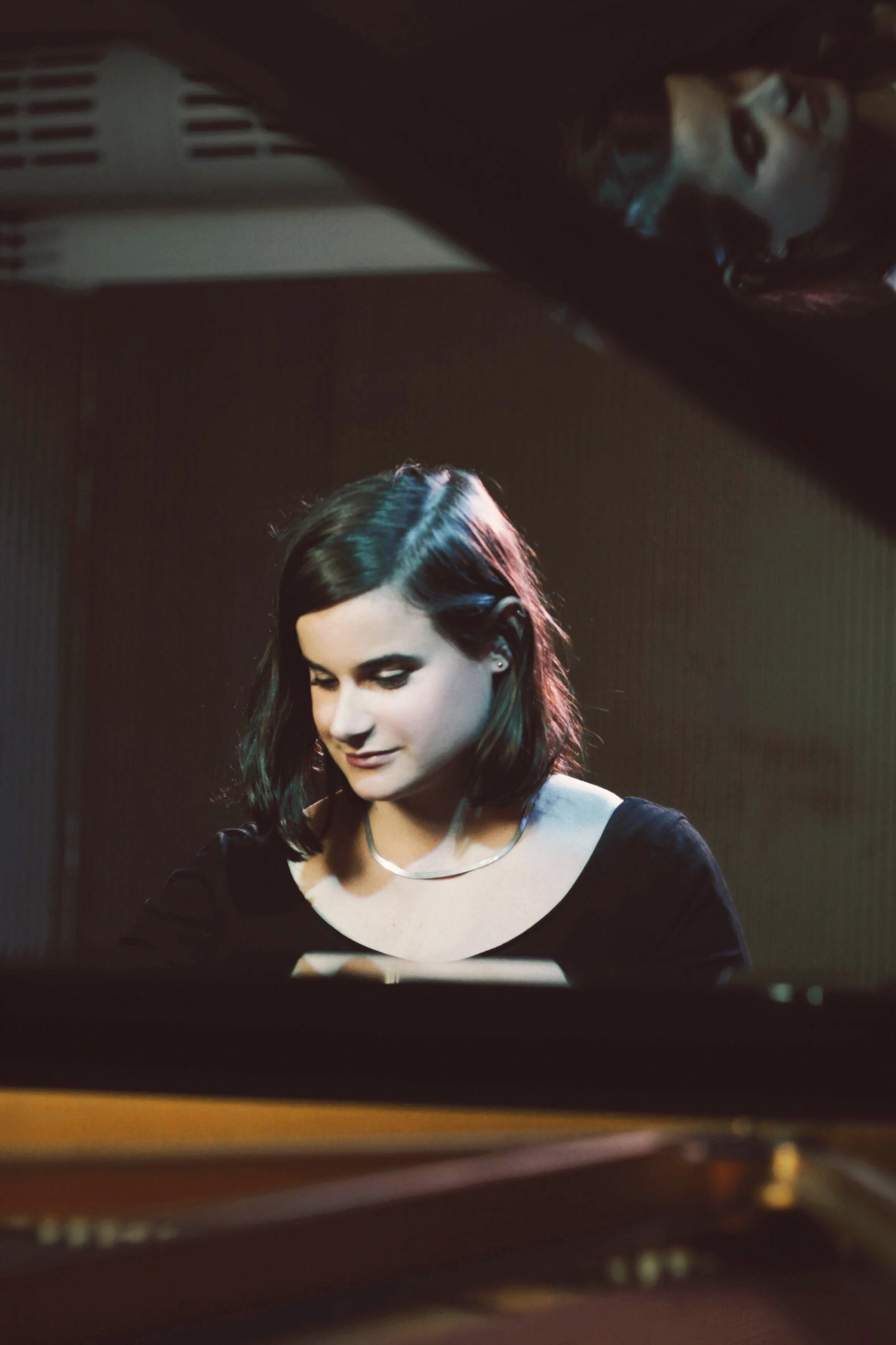 Une jeune femme avec des cheveux noirs et bouclés joue du piano, portant un vêtement noir et un collier, avec intérêt, dans une pièce sombre.