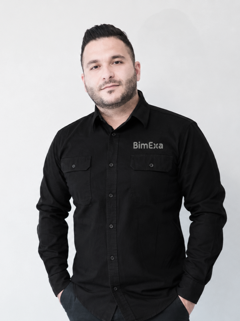 A man with dark hair and a beard, wearing a black shirt with the logo 'BimExa' on it, standing against a plain white wall.