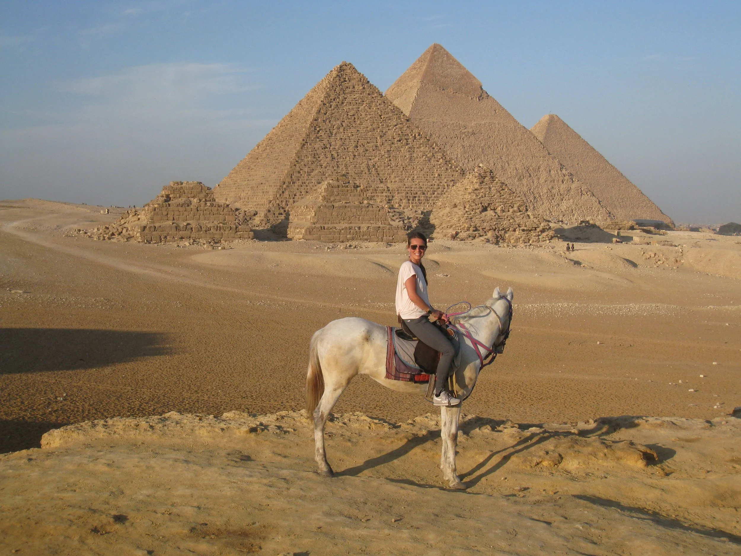 Woman riding a white horse in front of the pyramids of Giza in Egypt under a blue sky.