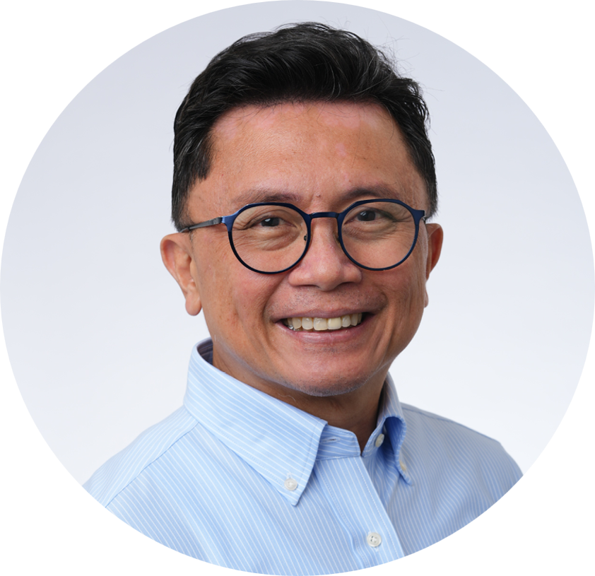 A smiling man with black hair, wearing glasses and a light blue striped button-up shirt, against a plain light background.