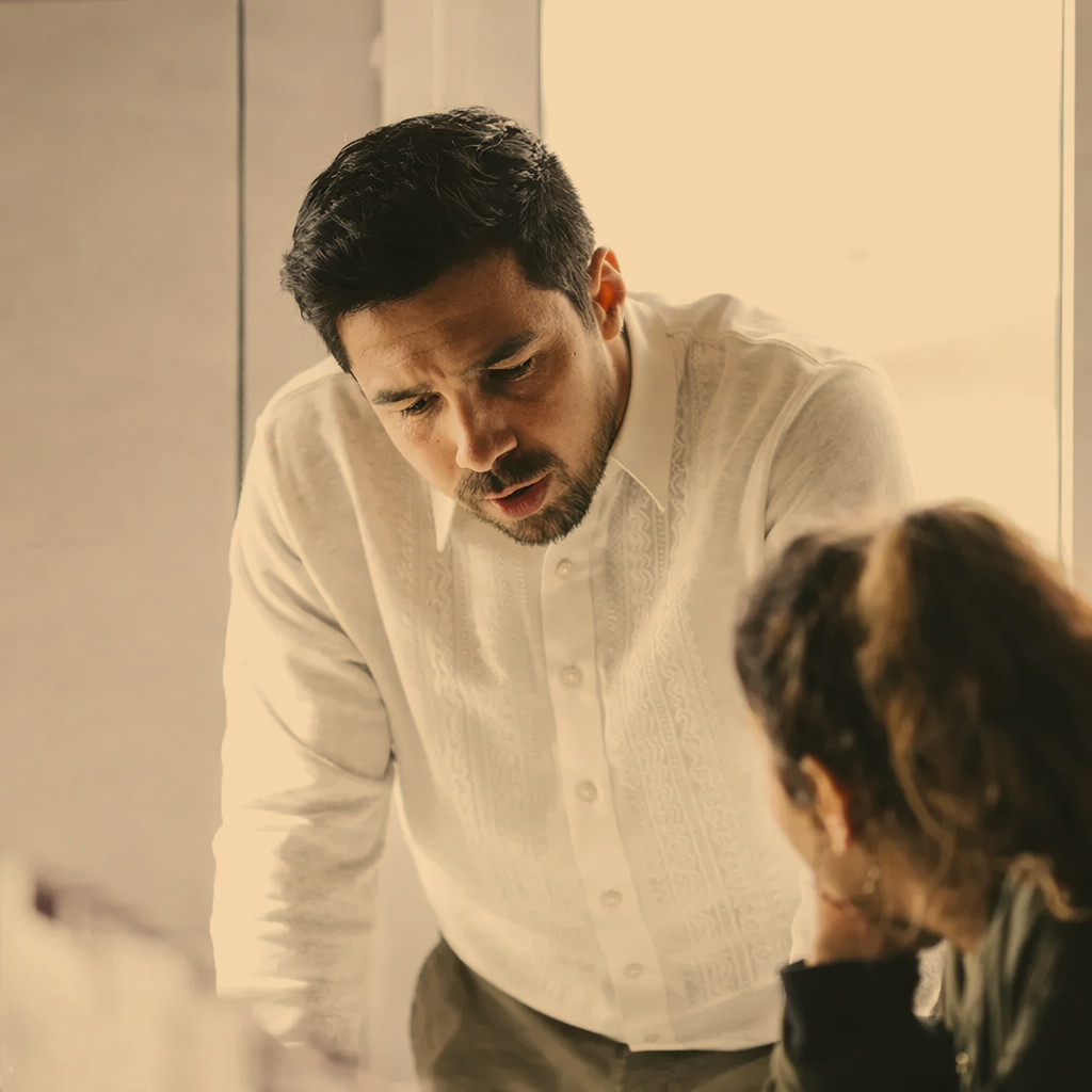 A man working with a woman who is seated to solve a business problem, with the woman's head resting on her hand, in an indoor setting with neutral-colored walls.
