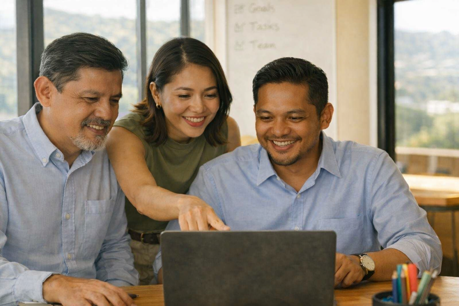 Three colleagues working together looking at a laptop screen and smiling in a bright office.