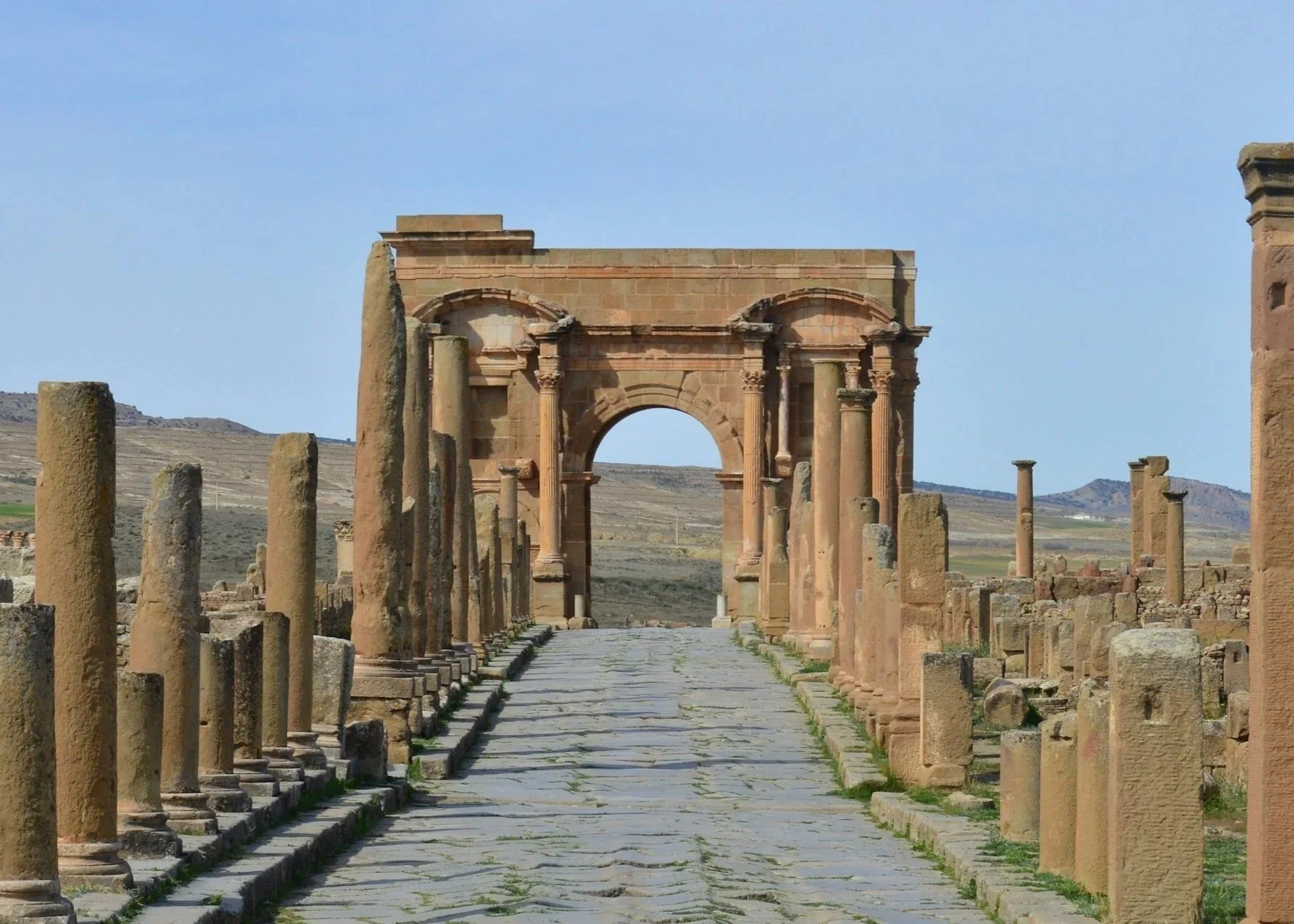 Ancient Roman ruins with a stone archway in a desert landscape, surrounded by columns and paved stone pathway.