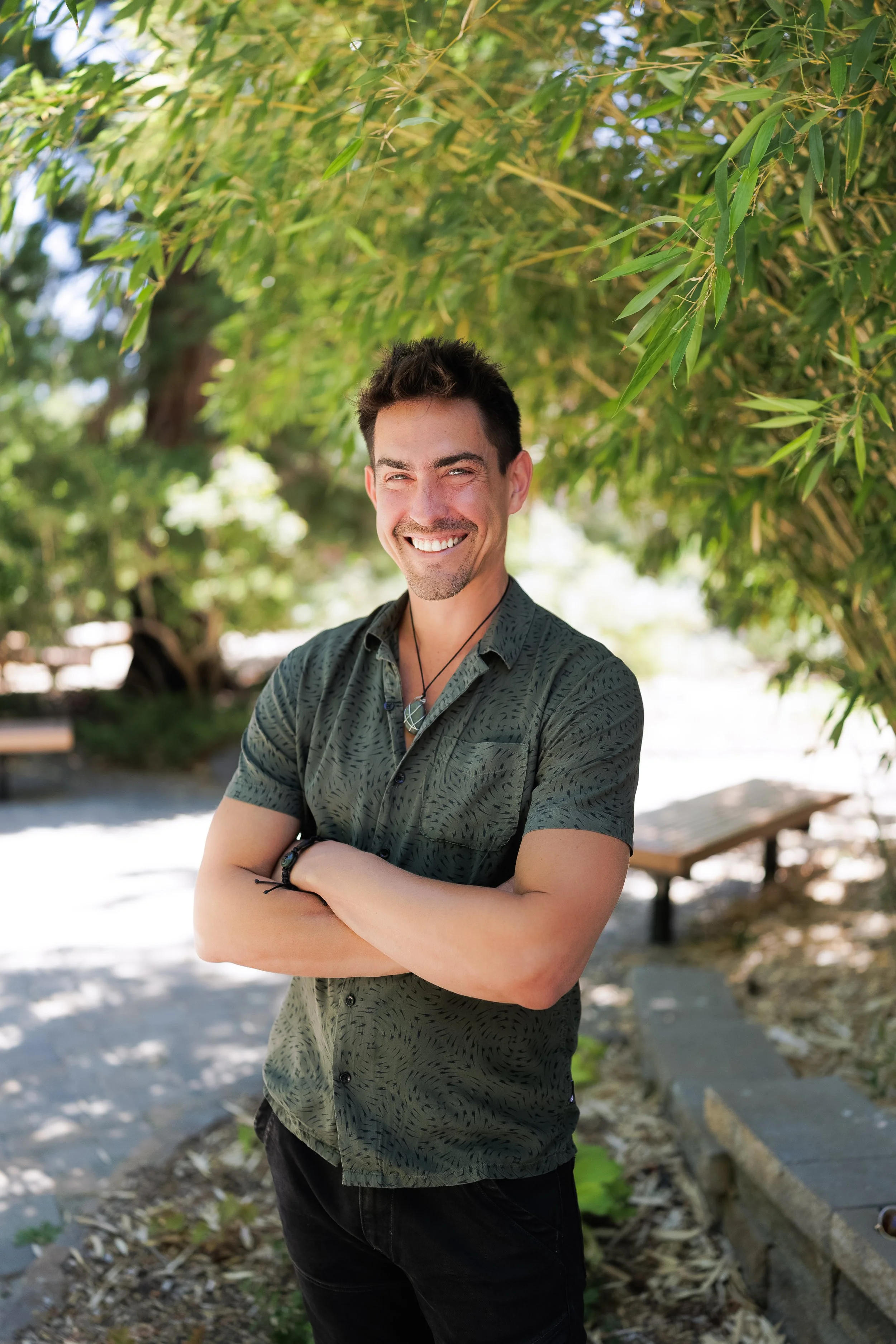A smiling young man with short dark hair, wearing a green patterned short-sleeve button-up shirt, stands outdoors under green leafy trees with his arms crossed in front of him.