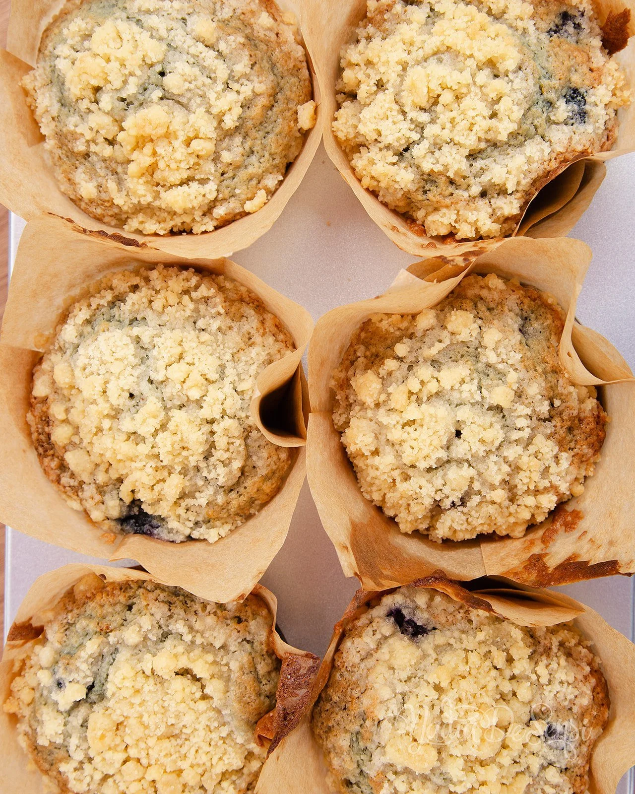 Six blueberry muffins with crumbly topping in parchment paper cups.