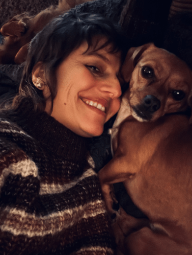 Eine Frau mit dunkelbraunem Haar und einem Hund, beide liegen eng zusammen und lächeln.