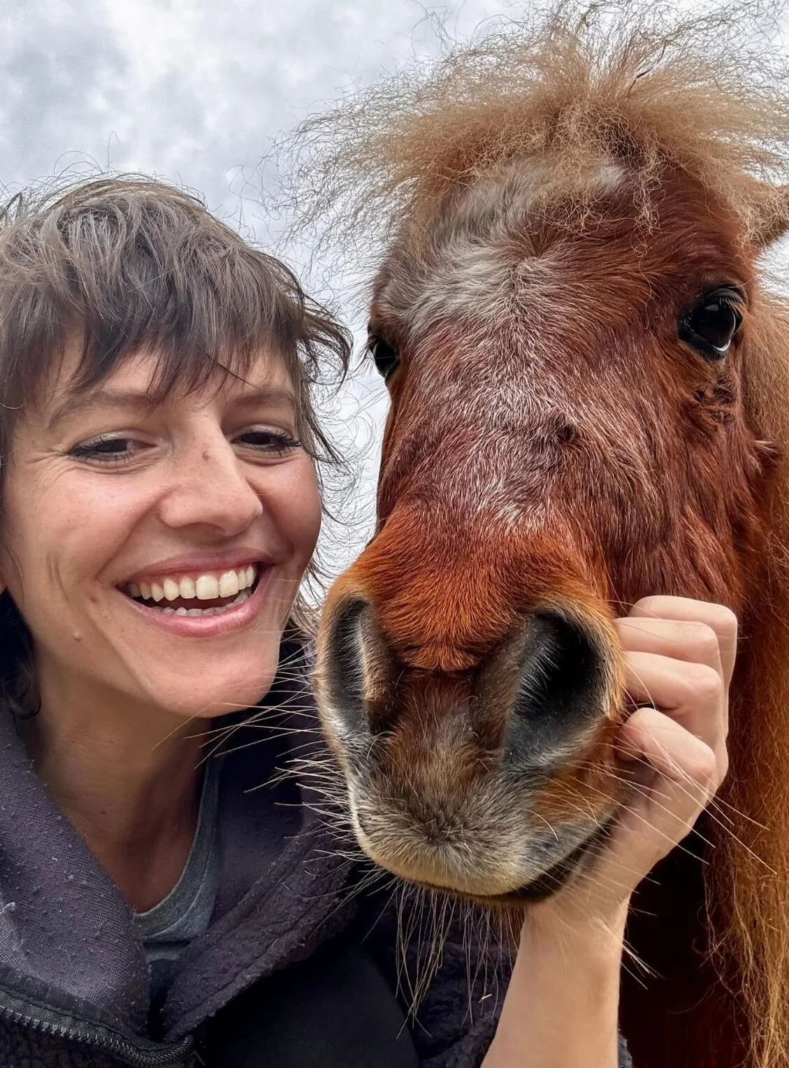 Lächelnde Frau mit kurz braunen Haaren, hält ein braunes Pferd an der Schnauze, beide schauen in die Kamera, im Hintergrund Himmel mit Wolken.
