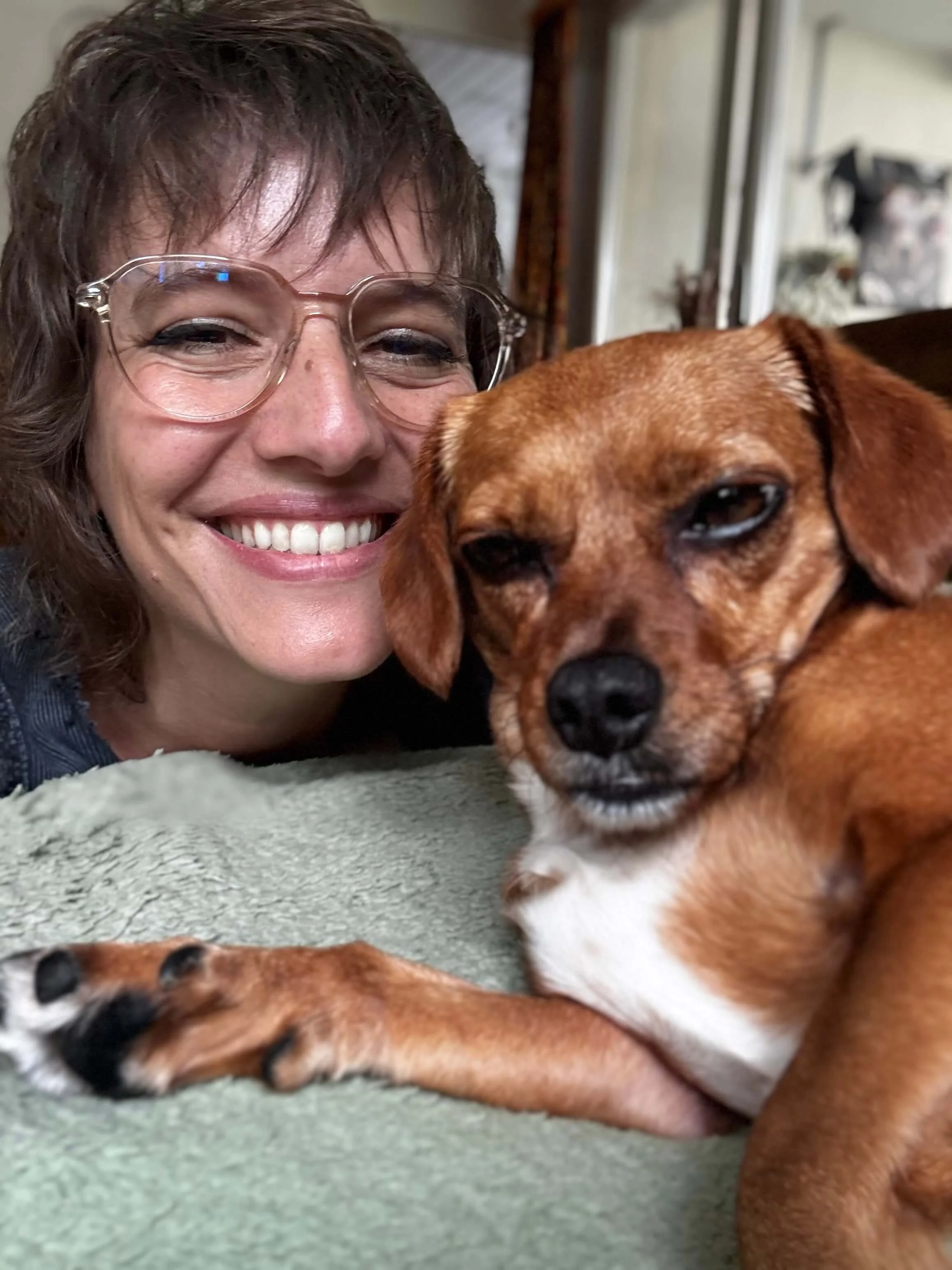 Frau mit Brille und braunem Haar lächelt, während sie einen braunen Hund streicht. Der Hund liegt auf einem weichen, grünen Oberflächen und schaut in die Kamera.