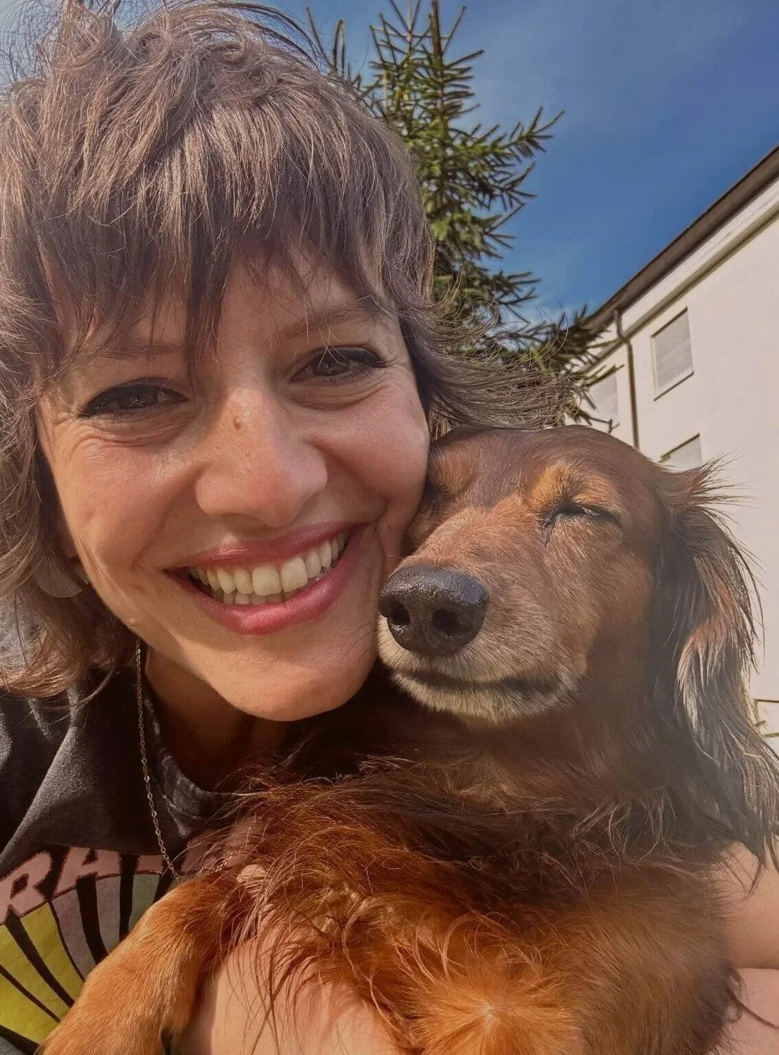 Vivian Vogel hält ihren entspannten Langhaardackel Max im Arm, draußen bei sonnigem Wetter, Baum und Haus im Hintergrund.
