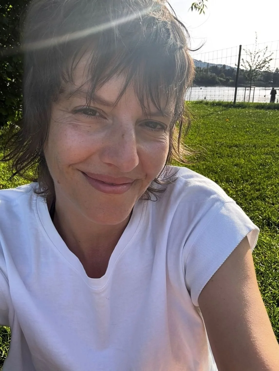 Ein lachender Mann mit braunen, nassen Haaren, trägt ein weißes T-Shirt, sitzt auf Gras im Freien, im Hintergrund ein Wasser und ein Zaun.