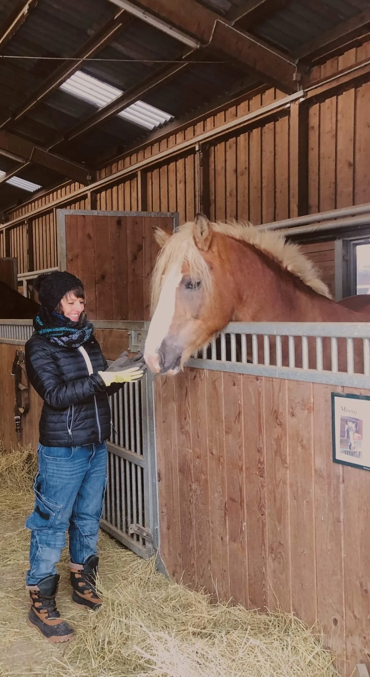 Eine Person in wetterfester Kleidung und Stiefeln streichelt ein hellbraunes Pferd in einem Pferdestall.