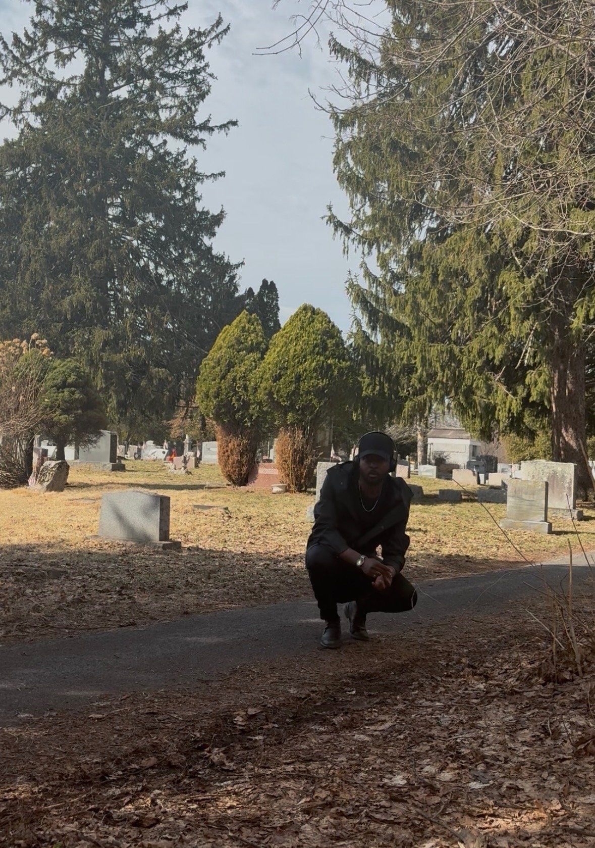 A person wearing dark clothing and a cap crouching on a pathway in a cemetery with tombstones, trees, and bushes in the background.