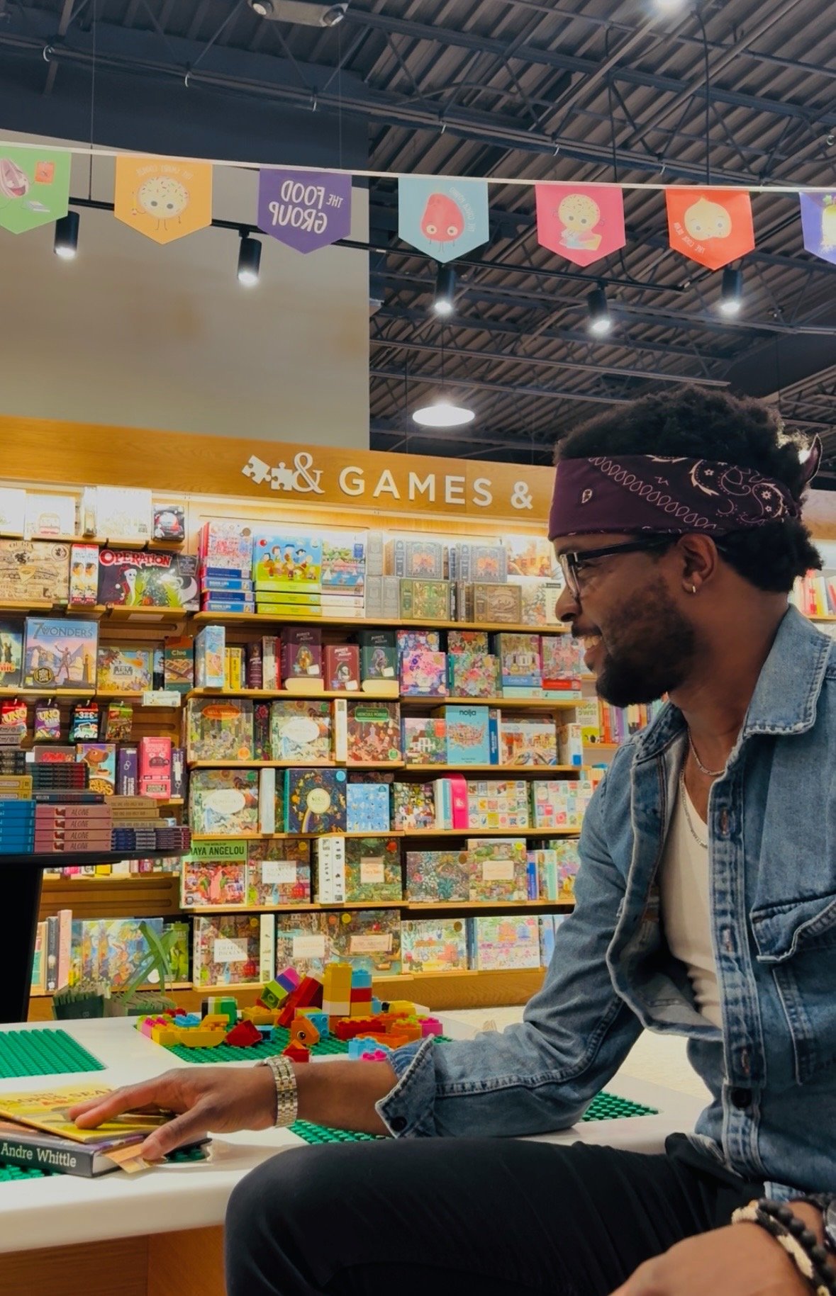 A man with curly hair wearing glasses, a purple bandana, a denim jacket, and a white shirt sitting at a table in a bookstore or toy store, playing with colorful Lego bricks. Shelves filled with books are visible in the background.