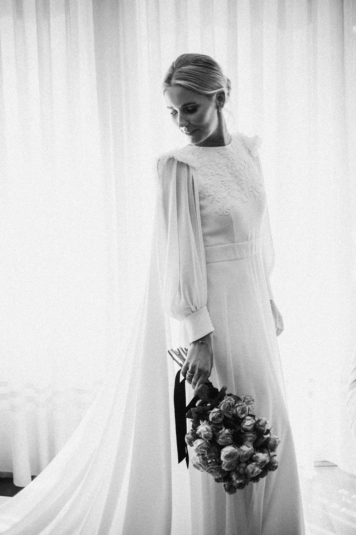 Novia vestido largo y mangas bombilla, sosteniendo un ramo de rosas, en un entorno de interior con cortinas lisas en Barcelona