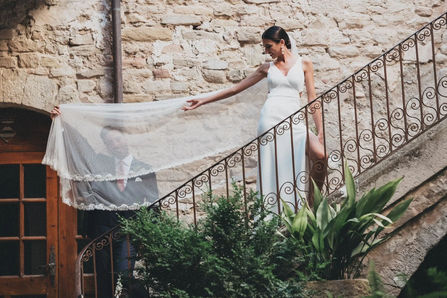 Una novia en vestido blanco en una escalera de hierro, con un fondo de pared de piedra en Castell de Tous