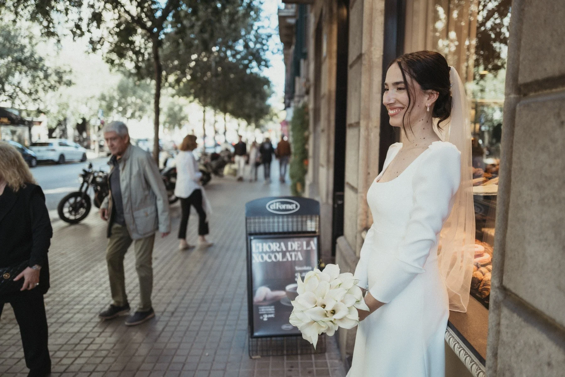Novia sonriendo, sosteniendo un ramo de flores blancas, de espaldas a la pared en la acera, con gente caminando y árboles al fondo