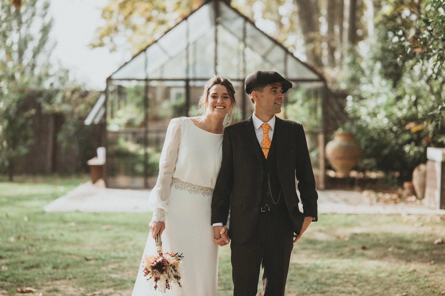 Pareja de boda caminando en un jardín con estructura de vidrio en el fondo, ambos sonriendo y sujetándose de las manos. Ella lleva vestido largo y brazos descubiertos, él lleva traje oscuro, camisa blanca y corbata naranja con puntos