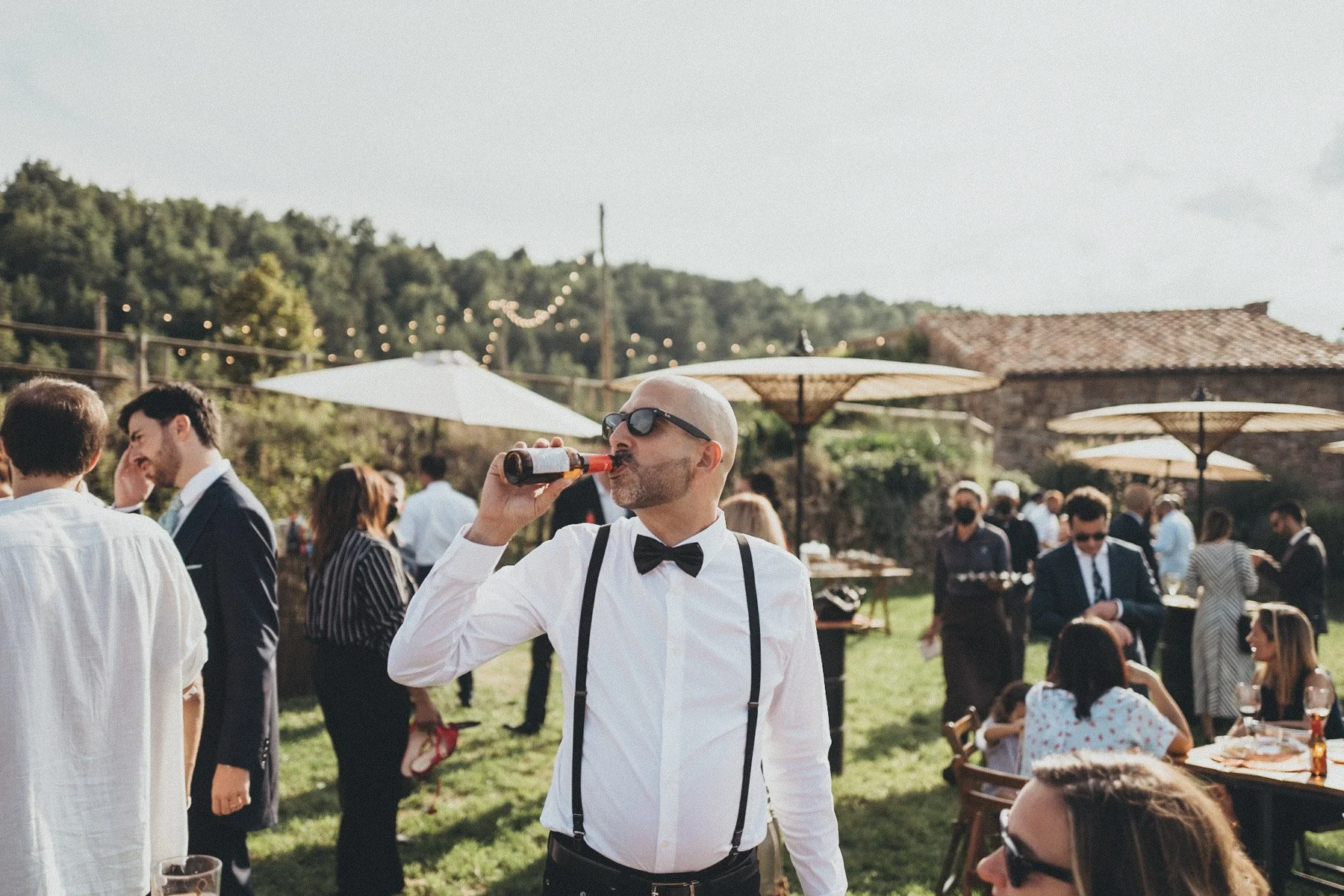 Hombre con gafas de sol, camisa blanca, pajarita, tirantes, que bebe una cerveza en una celebración al aire libre con varias personas en el fondo, algunas con trajes formales, en un ambiente rústico con sombrillas y luces decorativas.