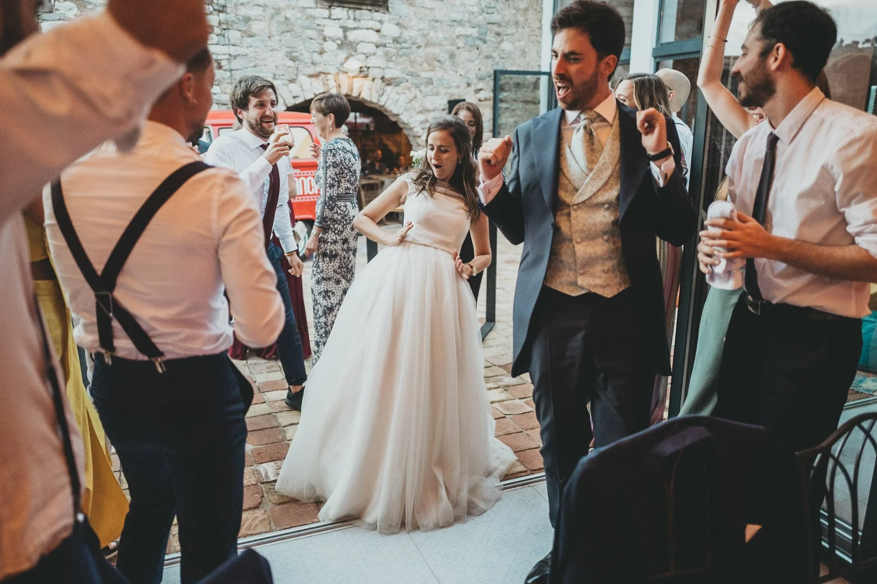 Grupo de personas bailando y disfrutando en una fiesta, con una novia en vestido de boda y un novio en traje, en un espacio con paredes de piedra y ventanas grandes.