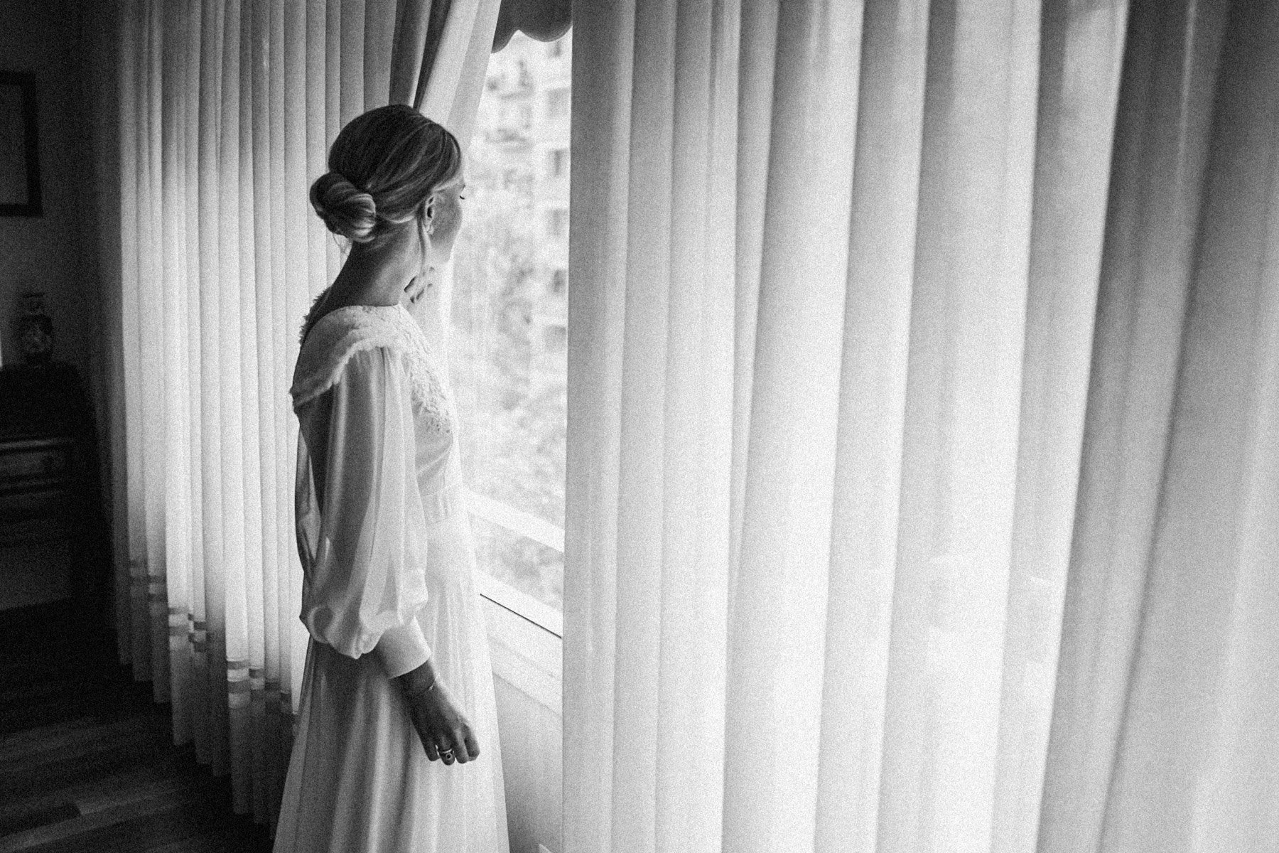 Mujer con vestido de novia de espaldas mirando por la ventana tras cortinas verticales.