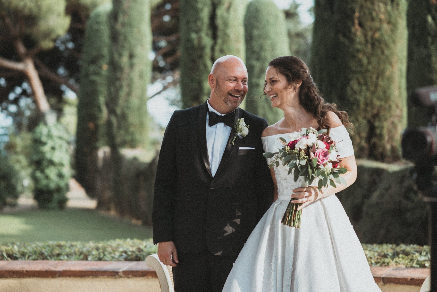 Una pareja de novios en un bosque, la mujer con vestido blanco y ramo de flores, el hombre con traje negro y pajarita, ambos sonriendo durante la sesión de fotos de Buenafortuna