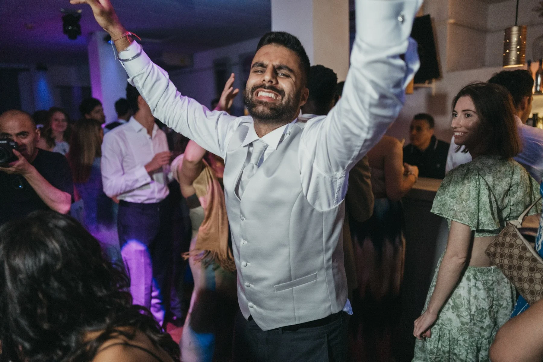 Un hombre con barba, vestido con una camisa blanca, bailando con los brazos levantados en una fiesta con otras personas en el fondo, algunas sonriendo y otras tomando fotos.