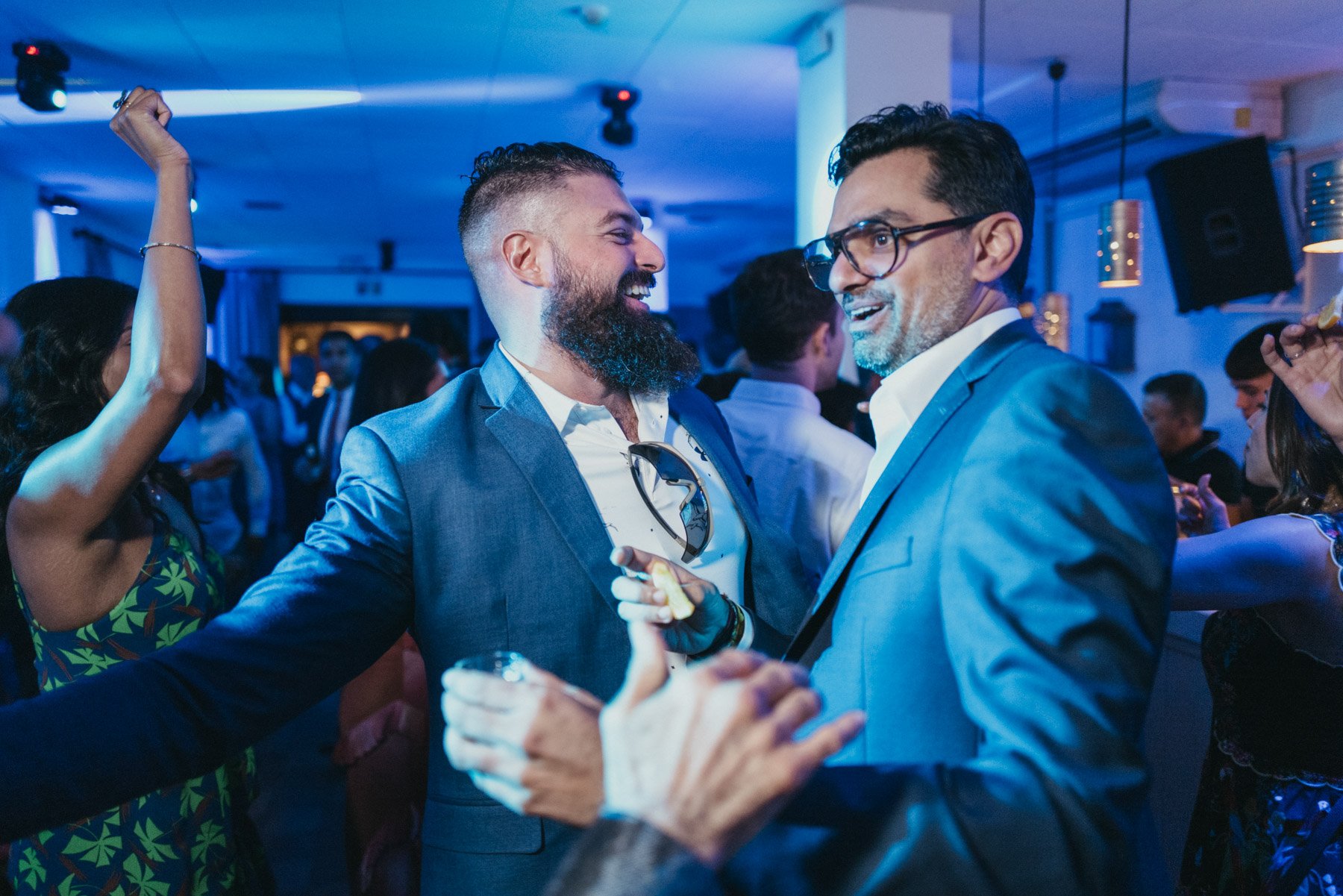 Dos hombres en trajes brindando y sonriendo en una fiesta en interiores con otras personas en el fondo