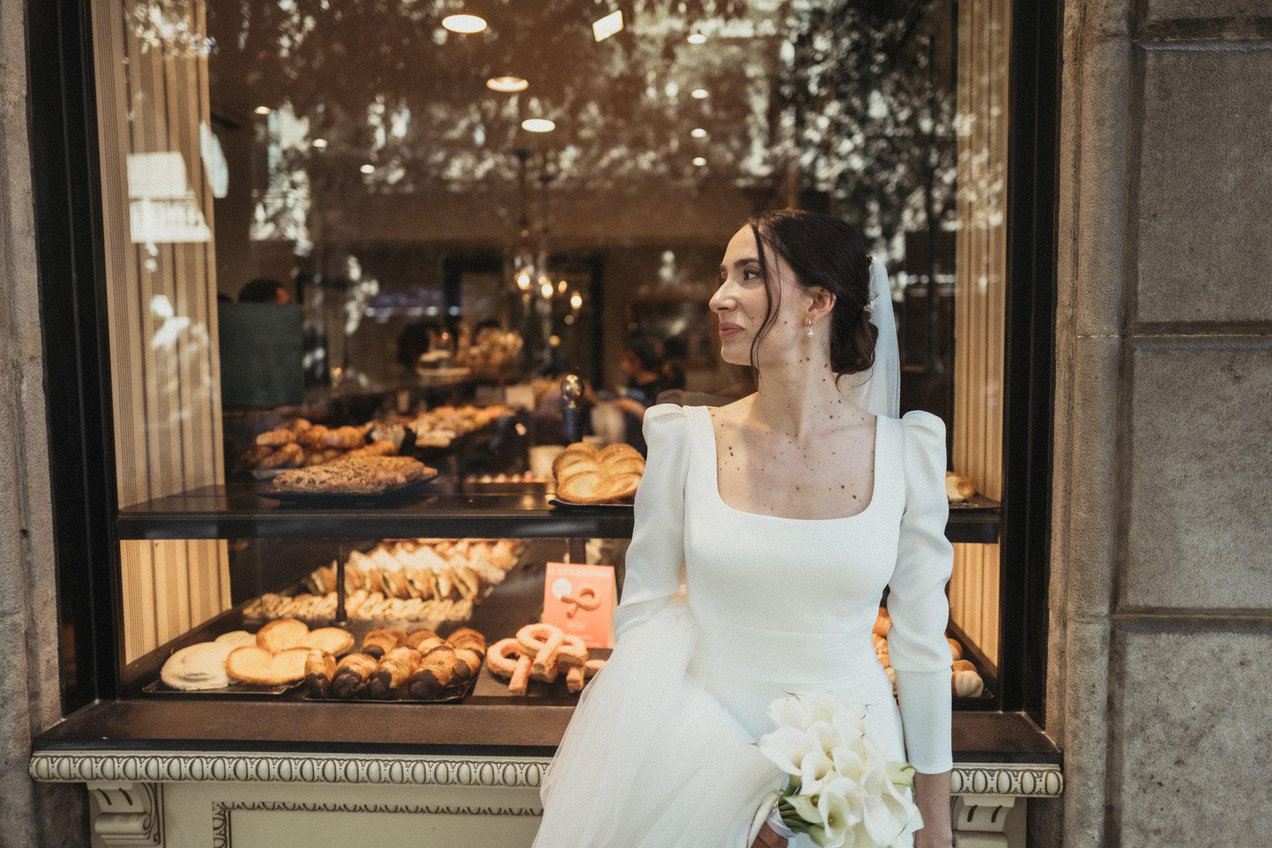 Joven mujer vestida con vestido blanco y velo de novia, sosteniendo un ramo de flores blancas, mirando hacia la izquierda frente a una vitrina con panes y pasteles en una cafetería o panadería.