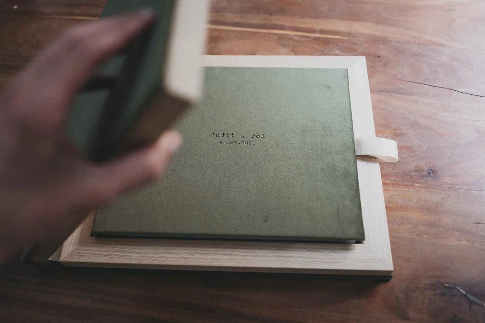 Álbum de boda color verde con texto en la portada sobre una mesa de madera descansando dentro de una caja con marco de madera. A green wedding album with embossed text on the cover, resting inside a wooden presentation box on a wooden table.