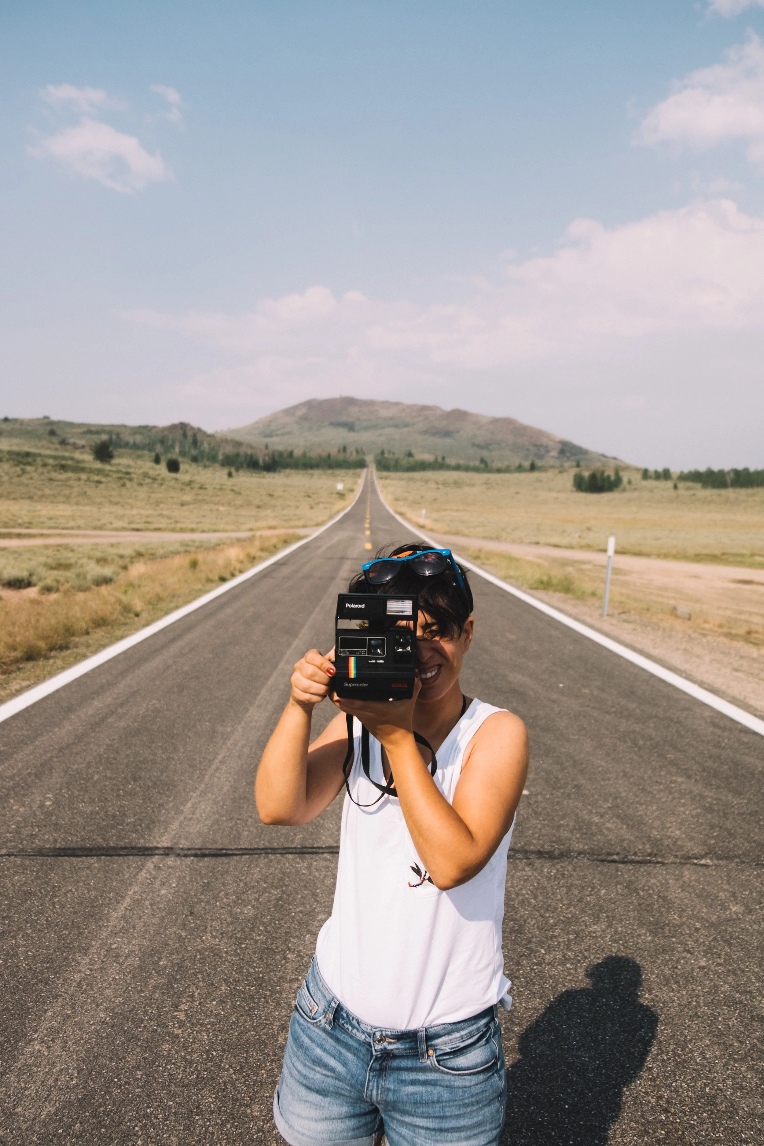 Mujer con camiseta blanca y jeans cortos tomando una foto en una carretera recta que se adentra en un paisaje de campo abierto con colinas en el fondo y un cielo despejado con algunas nubes.