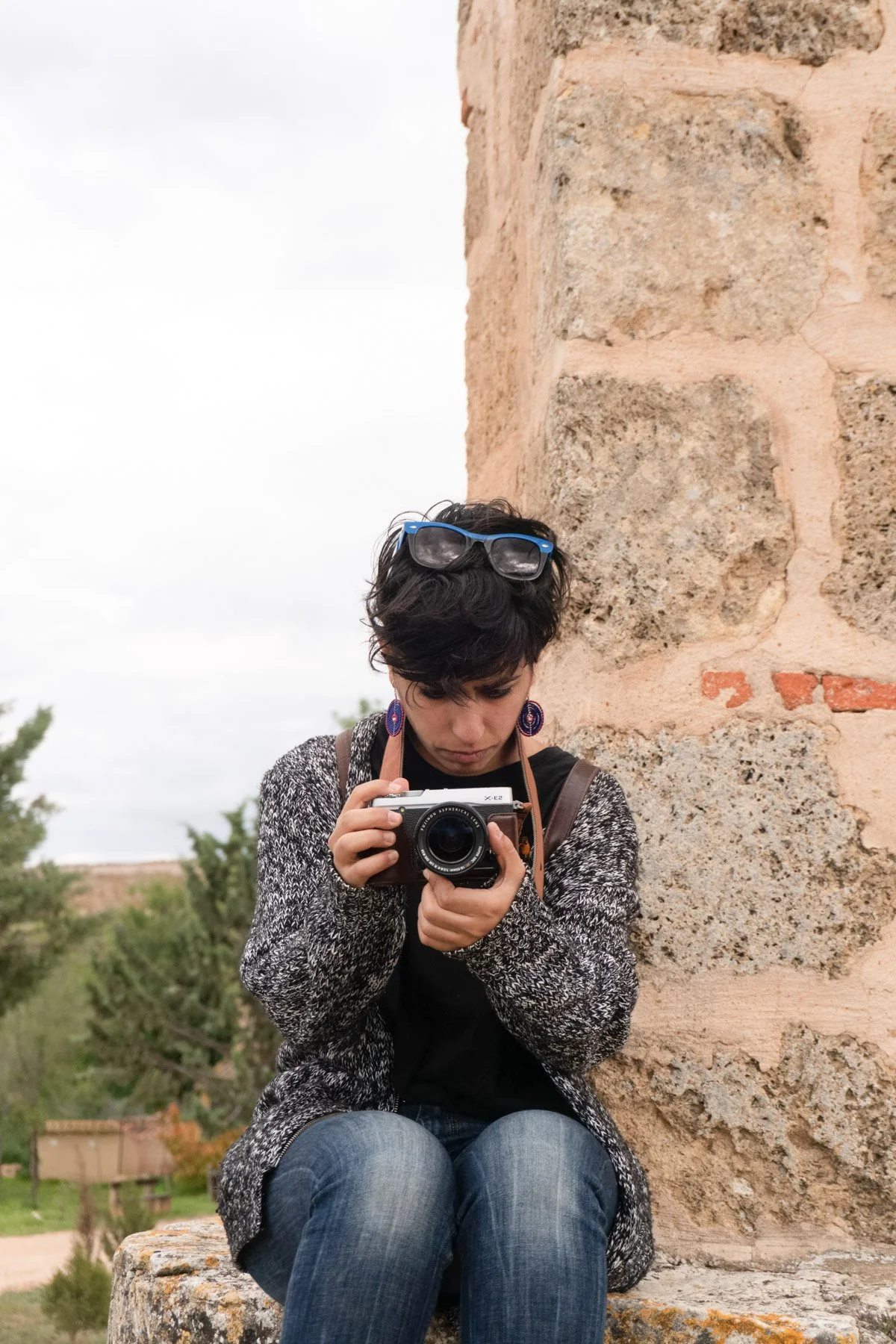 Videógrafa de Buenafortuna Studio está sosteniendo una cámara mientras mira hacia abajo. Ella está sentada en un muro de piedra junto a una pared de piedra en un entorno natural con árboles y un cielo nublado