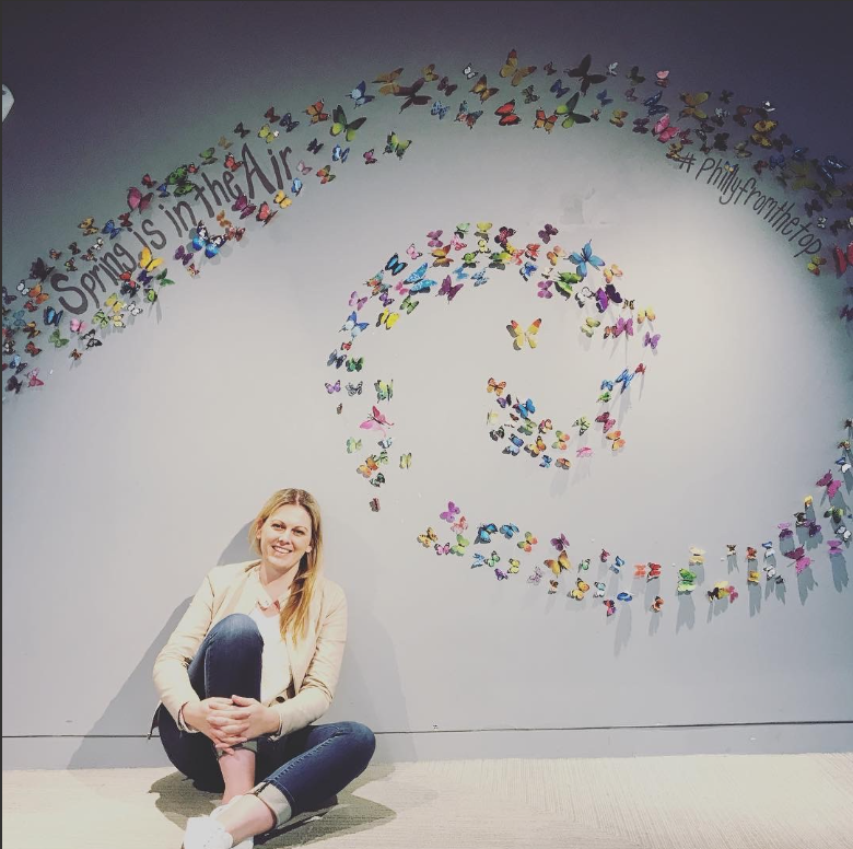 A smiling woman sitting on the floor in front of a white wall decorated with colorful butterflies and the phrases "Spring is in the Air" and "#Phillyfromthetop" written on it, in an exhibit or display.