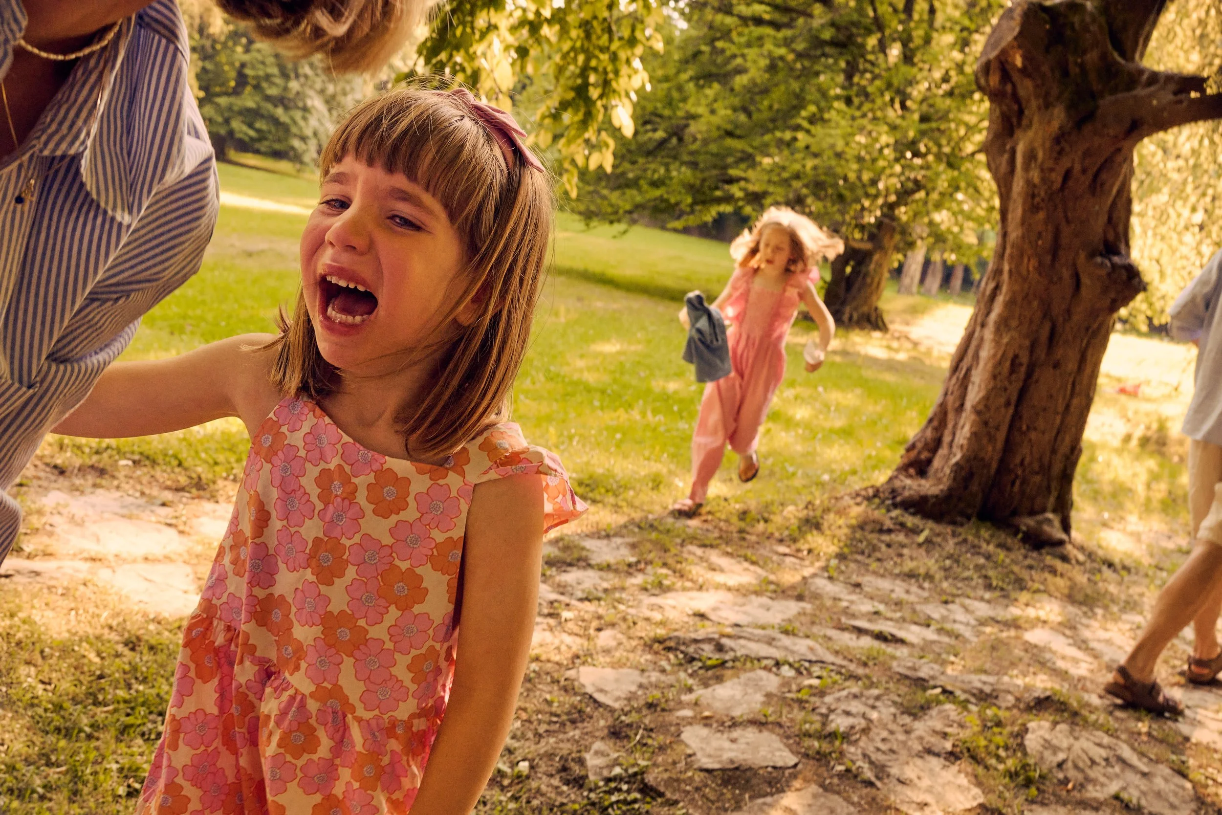 A young girl with a pink floral dress is outdoors, screaming or crying with her mouth open and eyes closed. Behind her, another girl in a pink outfit is running with her hair flowing. A woman with blond hair and a striped shirt is partially visible o