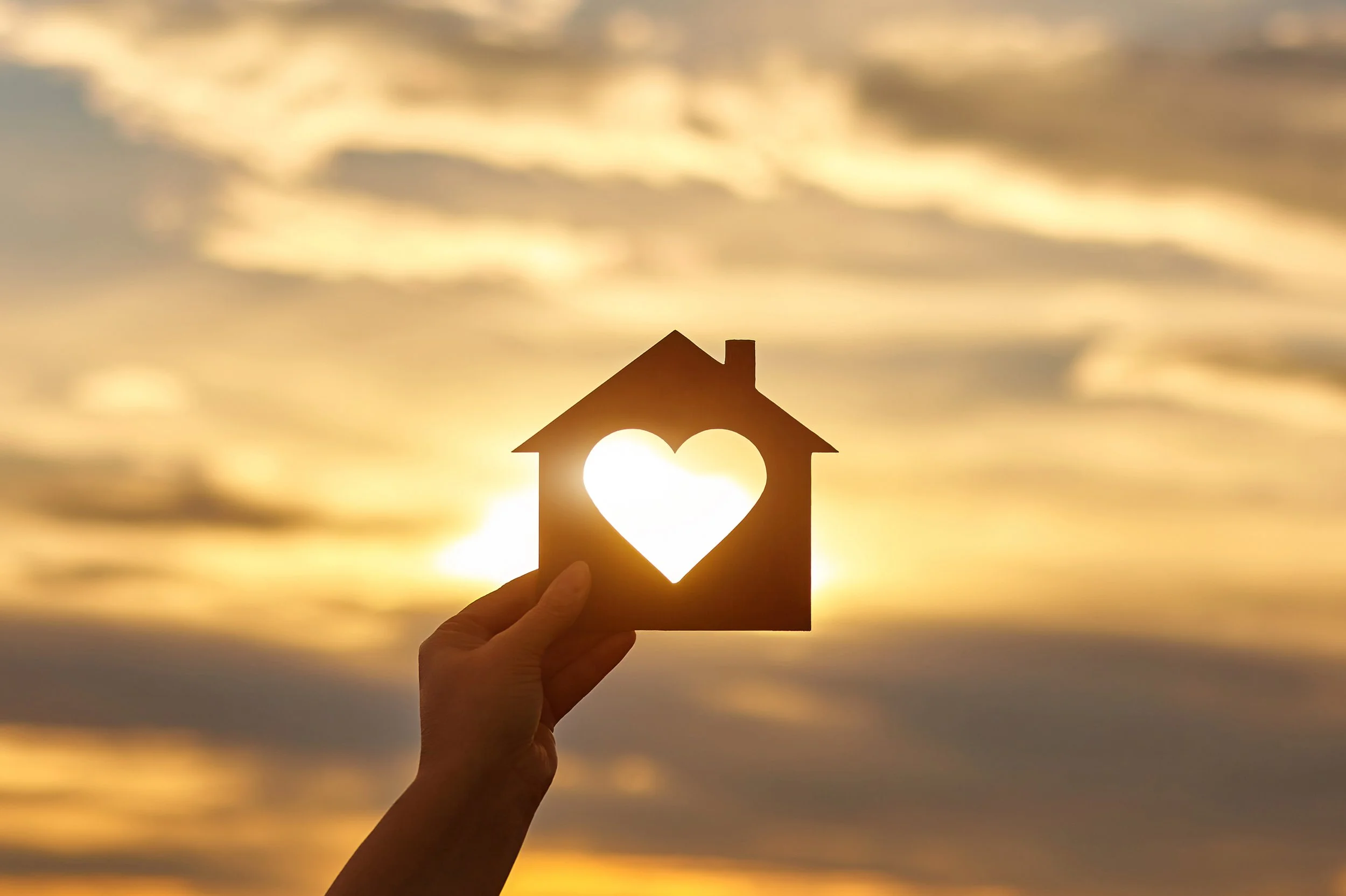 A person holding a paper house with a heart cutout in front of the setting sun, creating a silhouette with a golden sky and clouds.