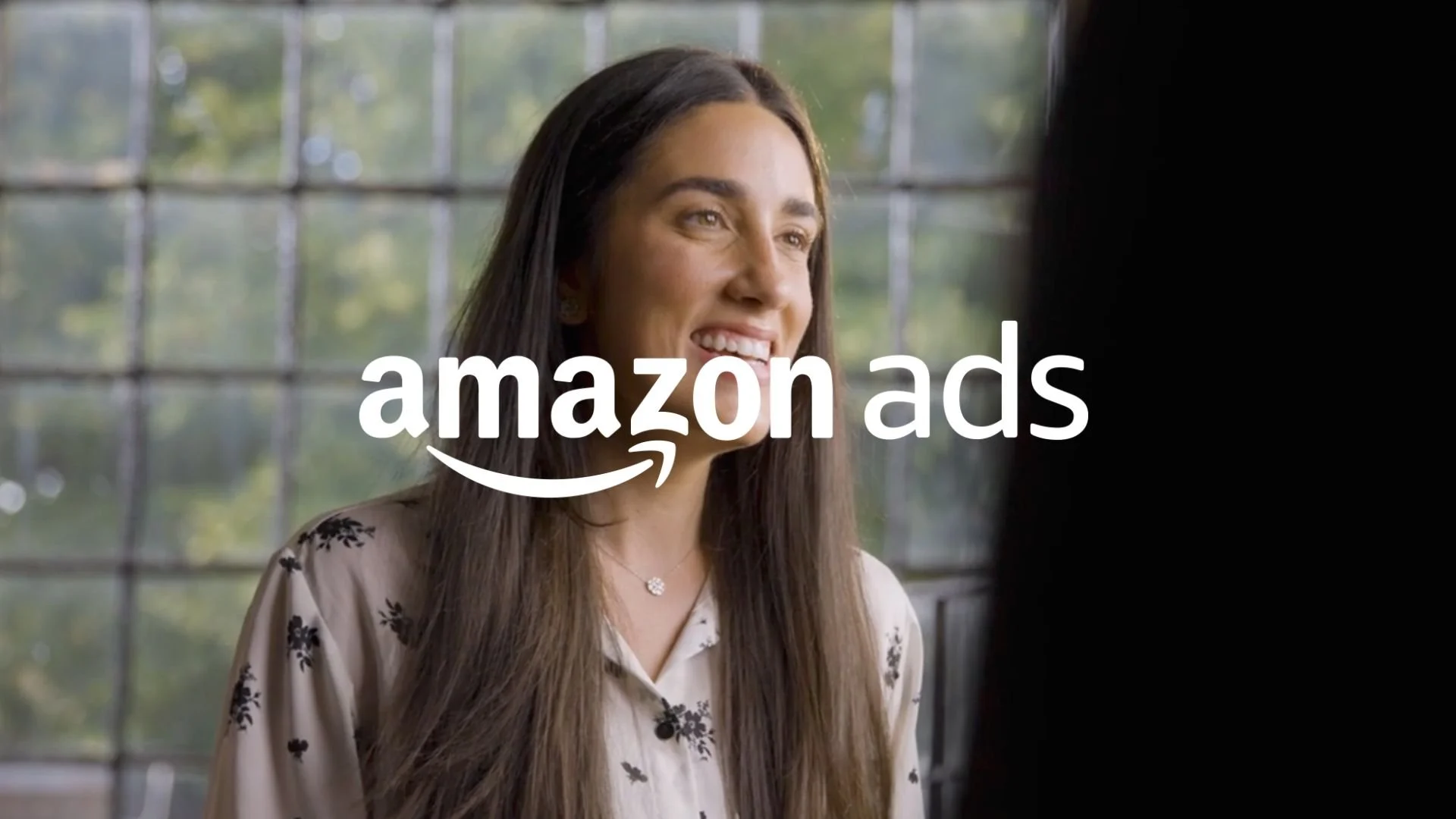 A woman with long brown hair smiling indoors with a window and trees in the background, overlaid with the text 'amazon ads'.