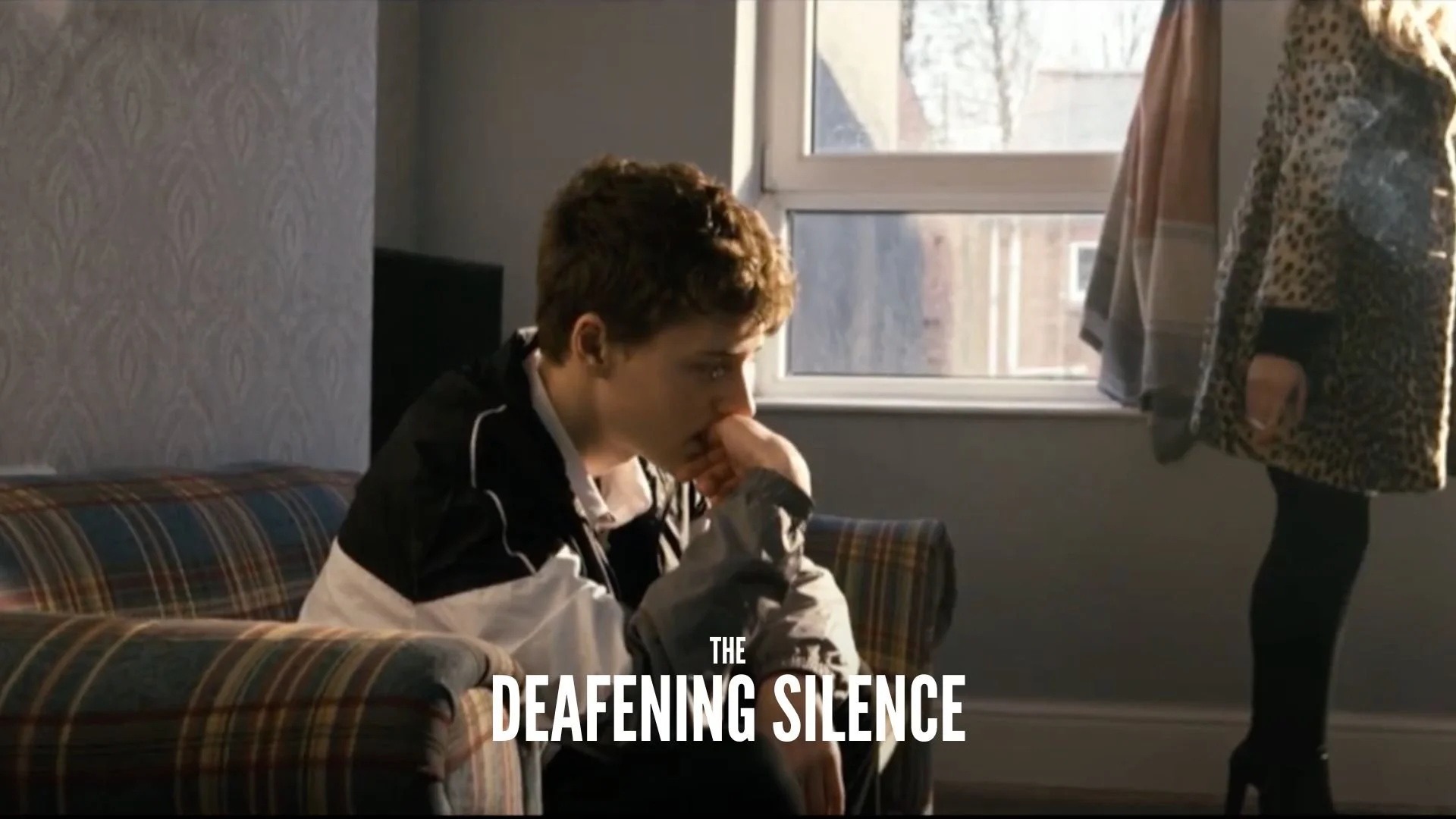 A young man with short curly hair sitting on a plaid couch with his hand covering his mouth, looking thoughtful. A woman standing next to him wearing a leopard print coat. Sunlight coming through the window makes the scene cozy. The text "The Deafeni
