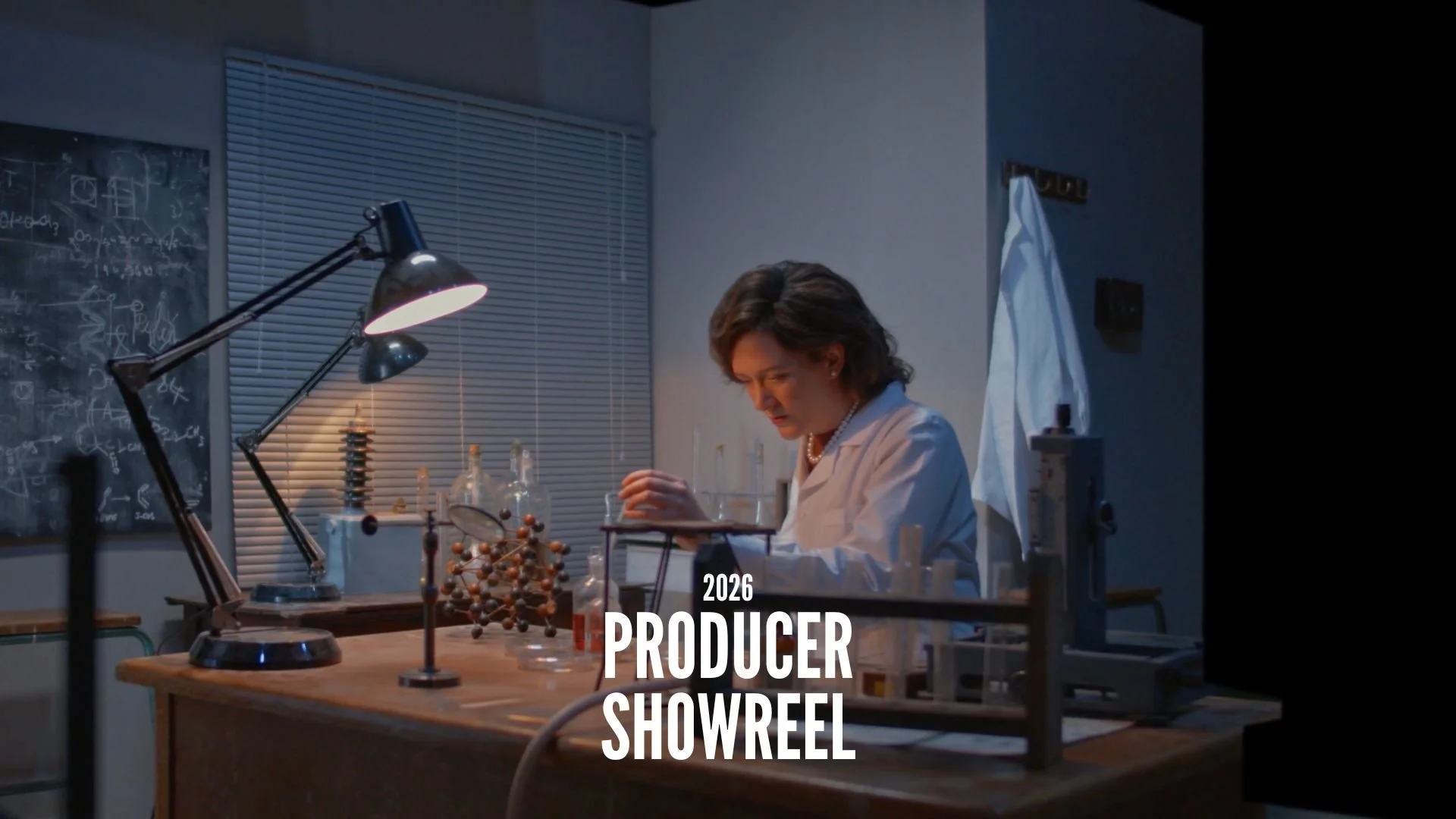 A woman working in a laboratory with scientific equipment, including test tubes, a molecular model, and a microscope, under a desk lamp, with a blackboard with scientific diagrams in the background. Text overlay: '2026 Producer Showreel'.
