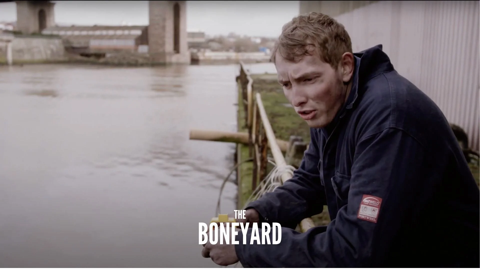 A man with disheveled hair and dirt on his face, wearing a dark jacket with a red logo patch, sits by the water near a railing under a bridge, with the words 'The Boneyard' overlaying the image.