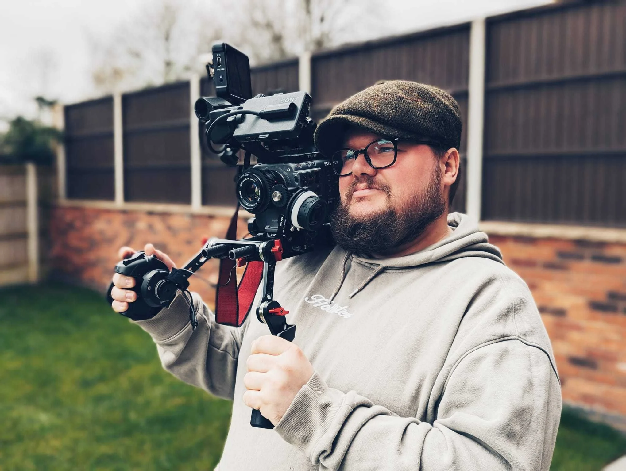 A man with glasses, a beard, and wearing a flat cap filming outdoors with a professional camera mounted on a stabilizer.