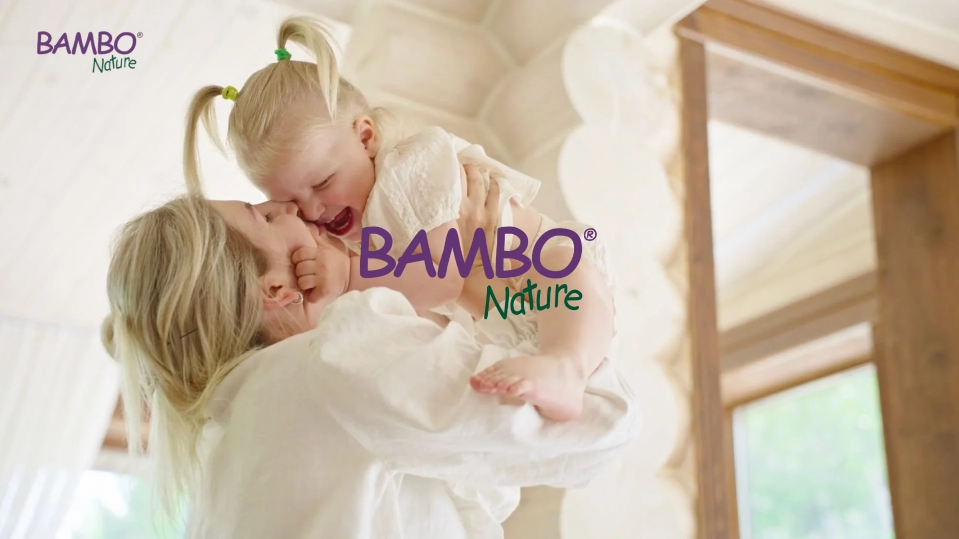 A woman and a young girl playing and smiling together inside a light-filled room with wooden beams.