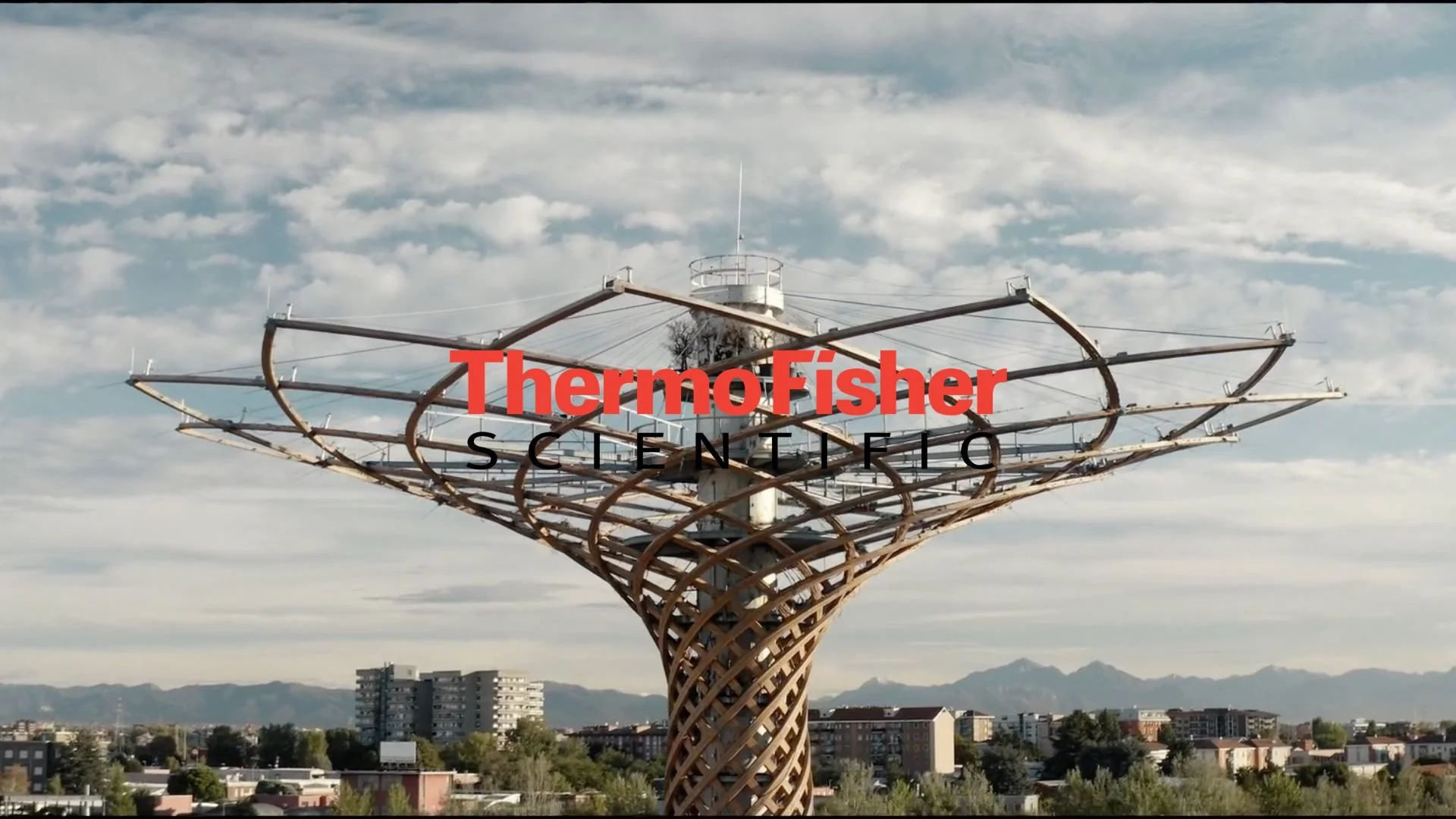 A tall, decorative tower with a lattice design, resembling a tree, under a cloudy sky with city buildings and mountains in the background. The words 'Thermo Fisher Scientific' are overlaid in red and black text.