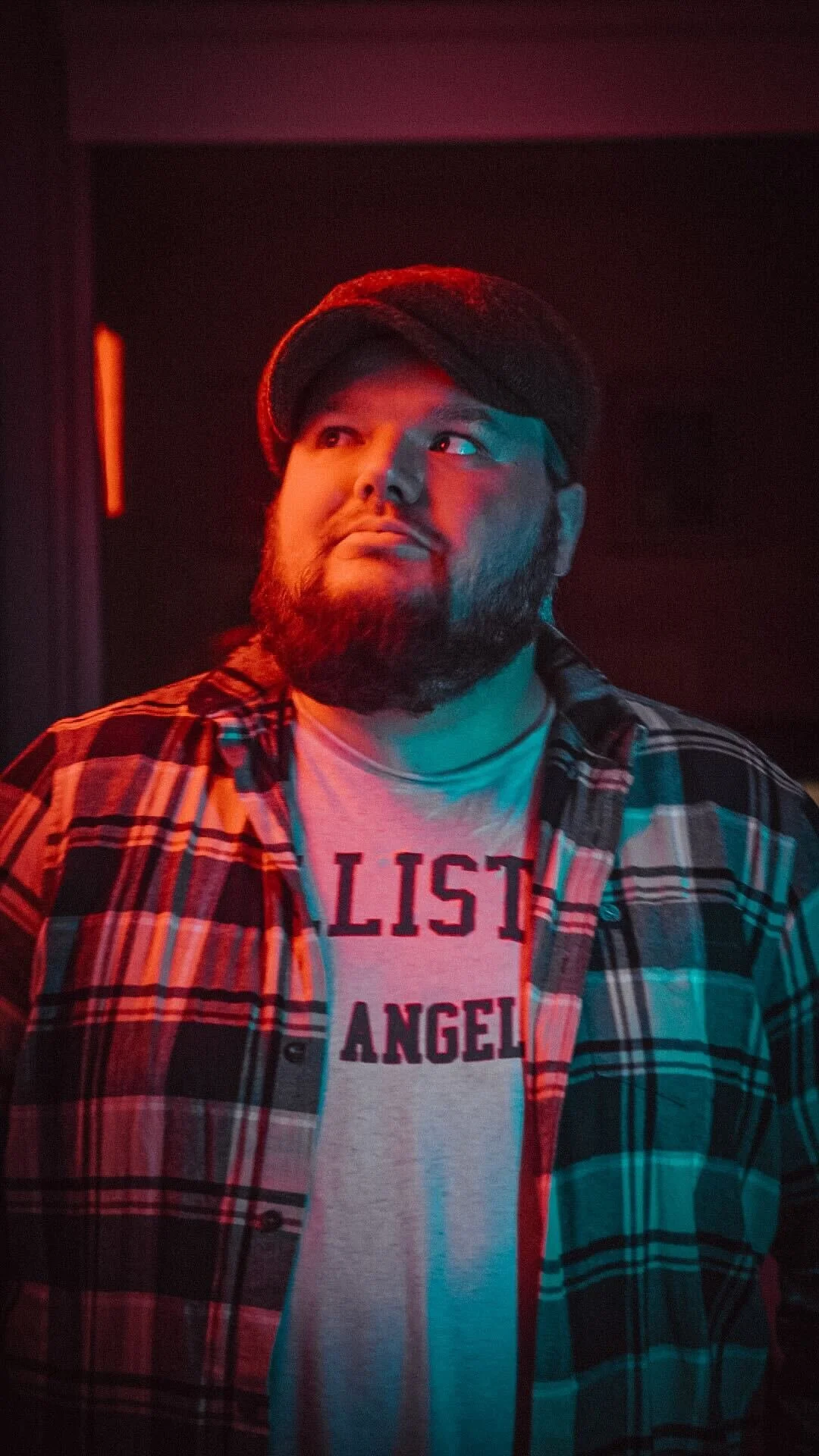 A man with a beard and mustache wearing a black flat cap and plaid shirt, with a white t-shirt underneath that says 'LIST ANGELES,' standing in a dimly lit setting with red and blue lighting.