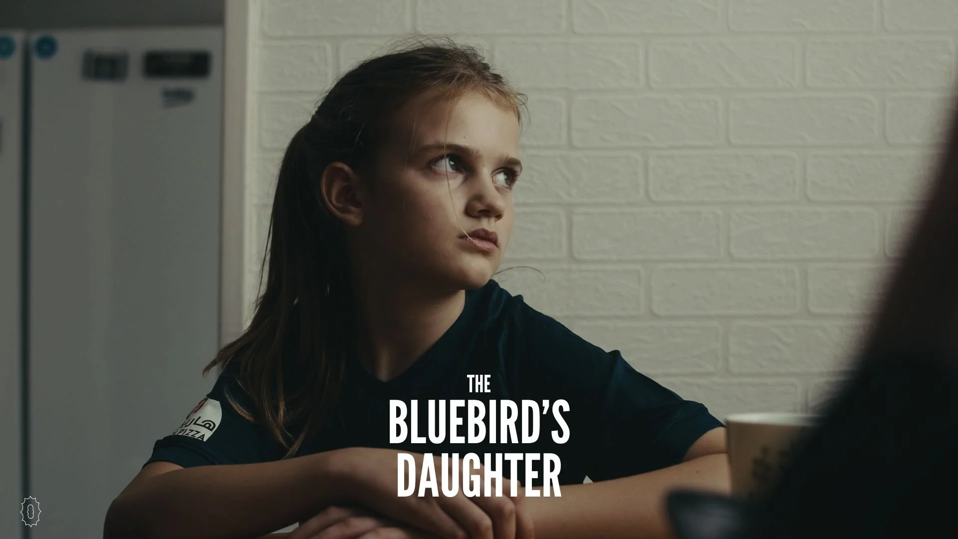 A young girl with long brown hair sitting at a table with a neutral expression, wearing a navy blue shirt, in front of a white brick wall, with a partial view of a coffee cup in the foreground. Text on the image reads "The Bluebird's Daughter."