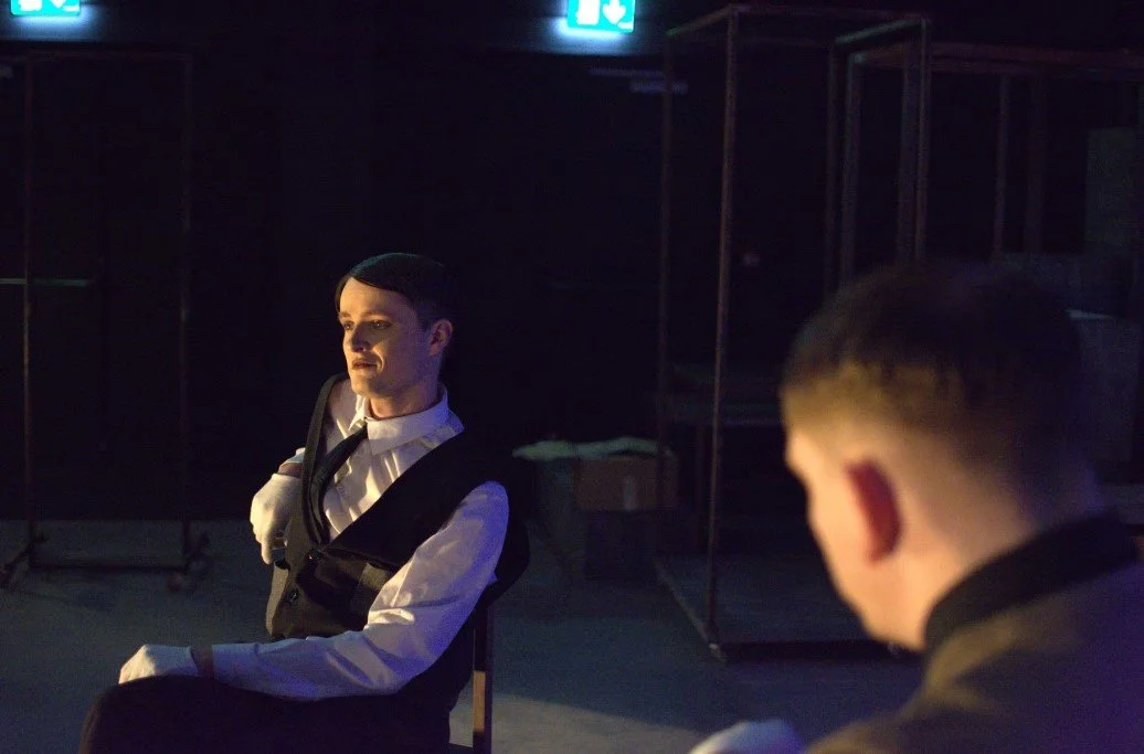 Two young men in formal attire sitting in a dimly lit room, with one facing forward and the other with his back to the camera.