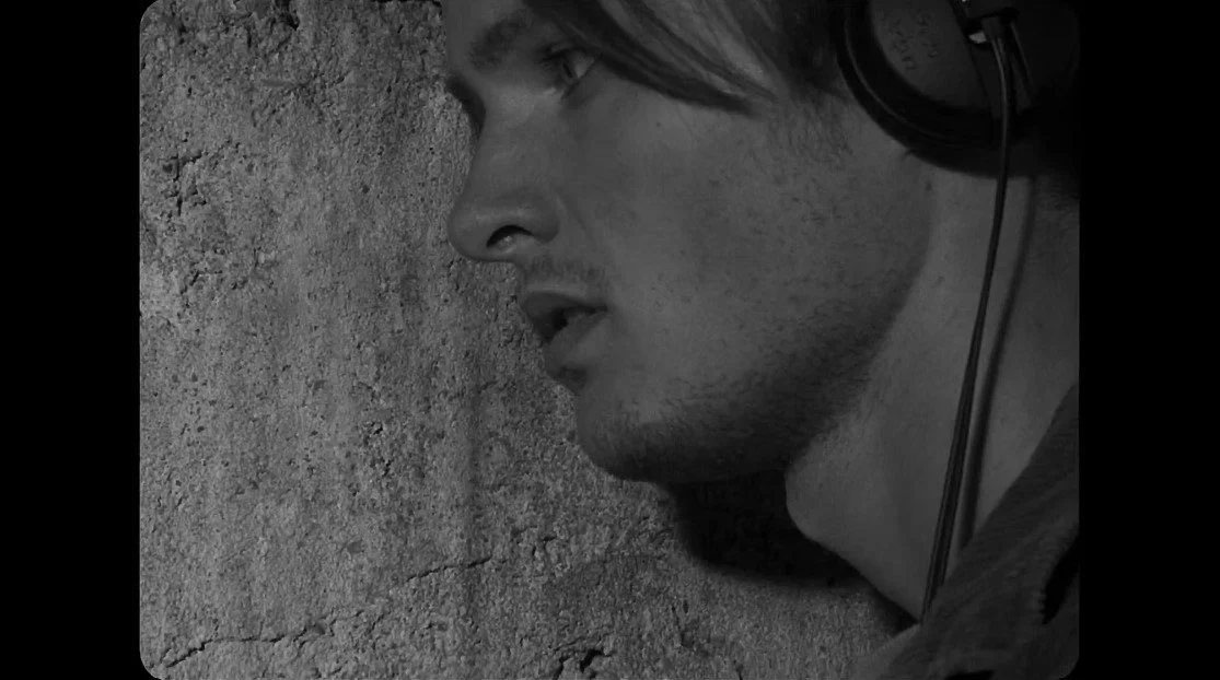 A young man with headphones resting his head against a textured wall, eyes closed or tired.