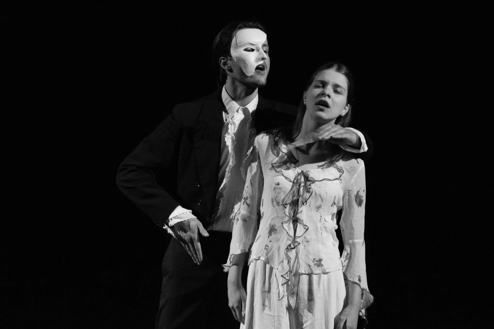 Black and white photo of two actors performing a dramatic scene on stage. The man is wearing a mask and speaking passionately, while the woman looks emotional.